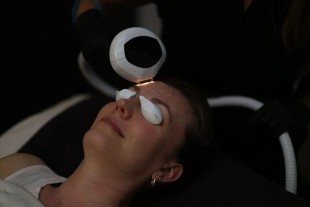Woman receiving a IPL facial treatment with a machine emitting light near her closed eyes in a dark spa or skincare clinic.