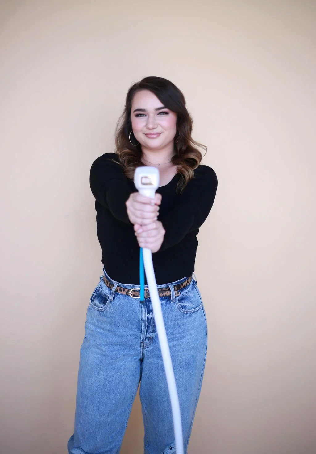 A young woman with wavy brown hair, makeup, and hoop earrings, smiling, holding a laser hair removal tool directed forward, standing against a plain beige wall, wearing a black sweater, blue jeans with a leopard print belt.