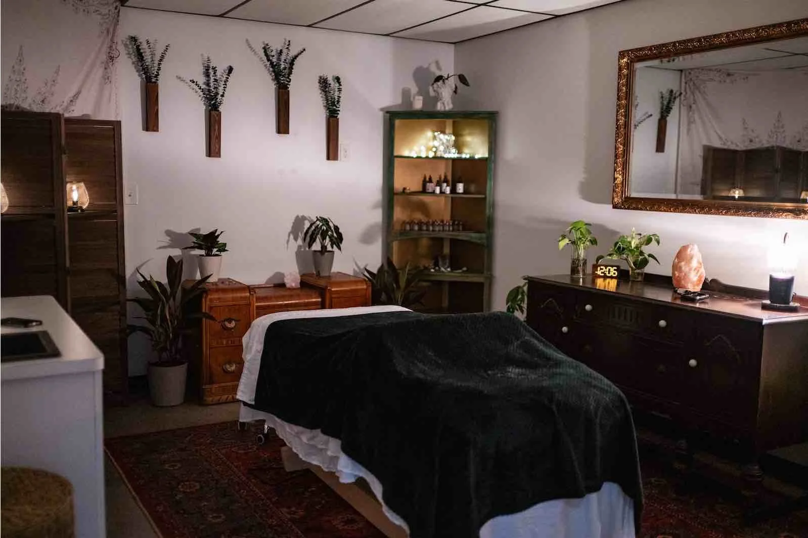 A cozy room with a bed covered in a black blanket and white sheets, surrounded by various plants, a large mirror, and wooden furniture, illuminated by soft lighting.