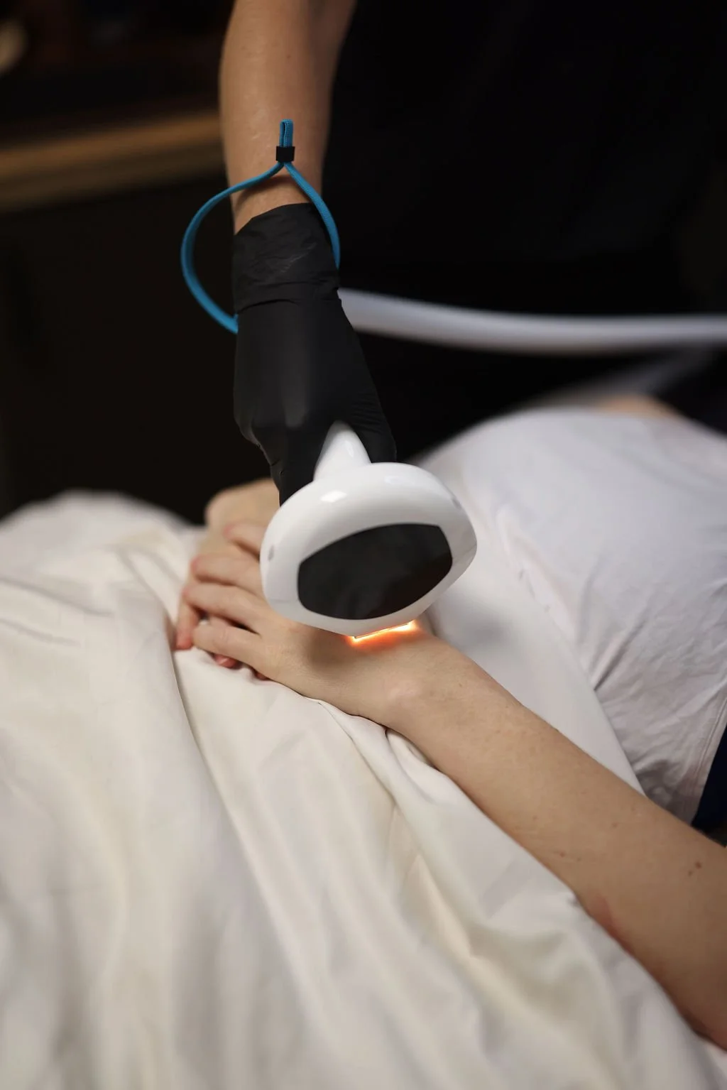 A person undergoing a medical or cosmetic procedure using a handheld device emitting red light, with their arm resting on a white sheet, wearing a black glove, and a blue wristband attached to their arm.