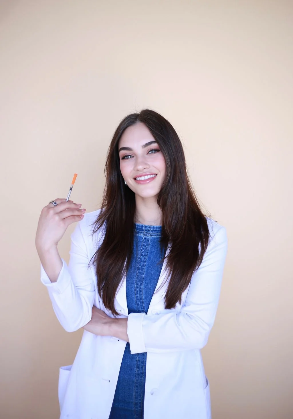 A smiling young woman in a white coat holding a syringe with an orange cap against a beige background.