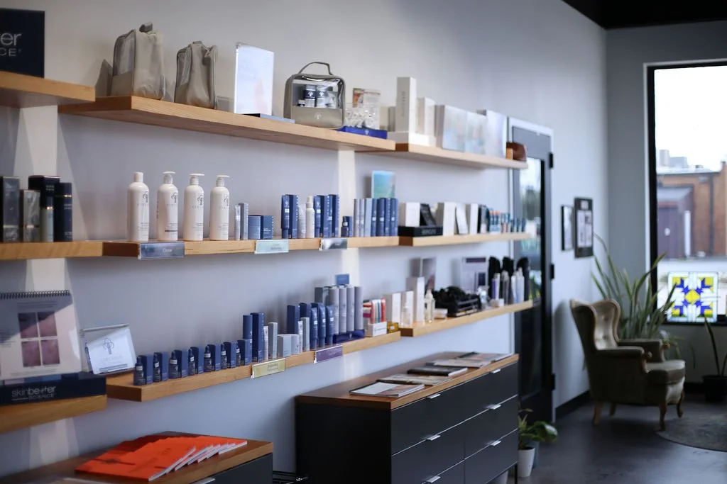 Interior of a Emily Anne Esthetics lobby with wooden shelves displaying skincare products, books, and small boxes. A comfortable seating area with chairs and a window with a colorful stained glass design.