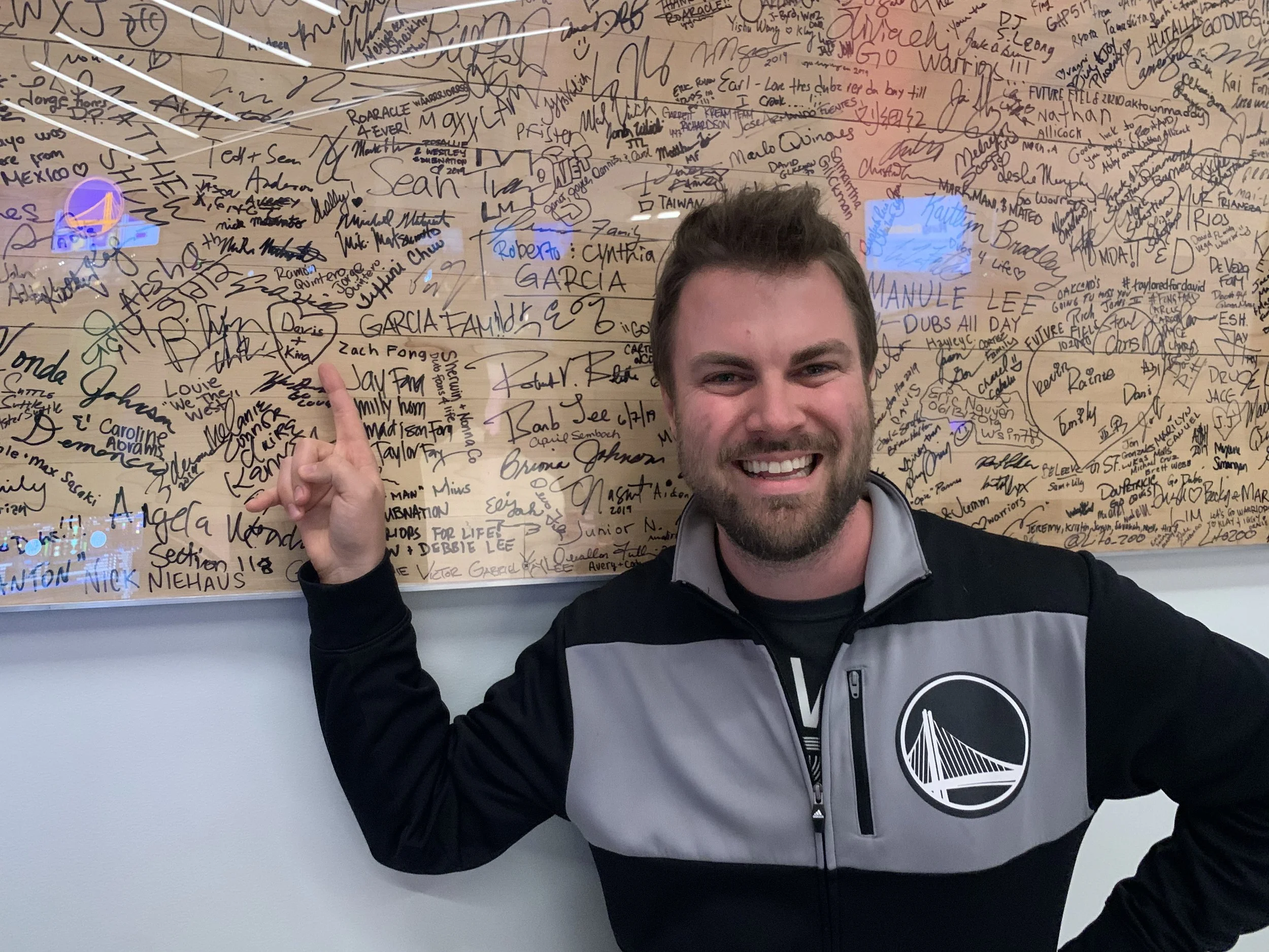A smiling man with a beard pointing at a wall covered in signatures and messages, with a digital clock displaying 4:12 and a blue sign with initials or numbers.