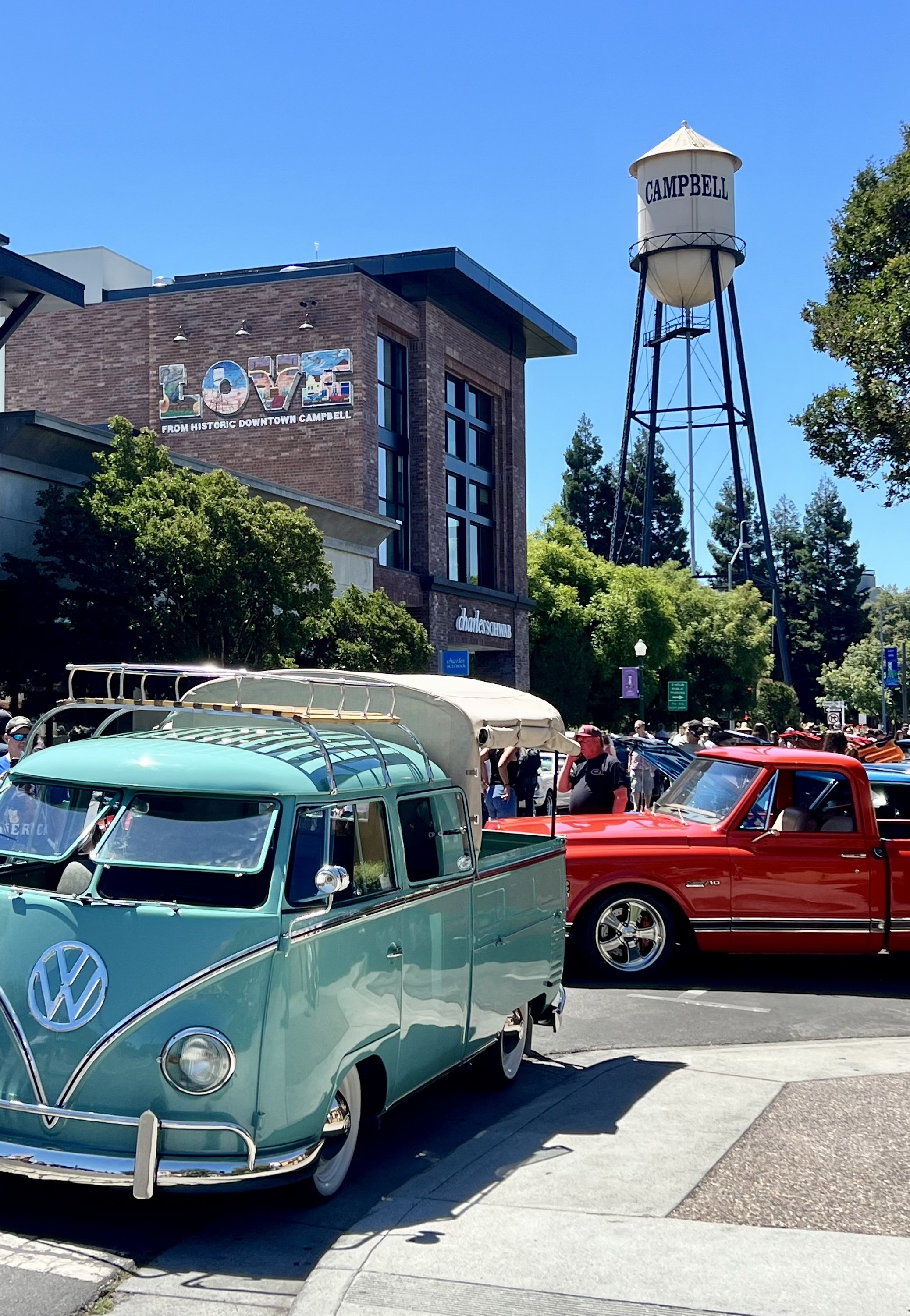 A vintage turquoise Volkswagen van and a red pickup truck parked on a street during a car show in Campbell, California. People are walking around, and there are trees and historic buildings in the background.