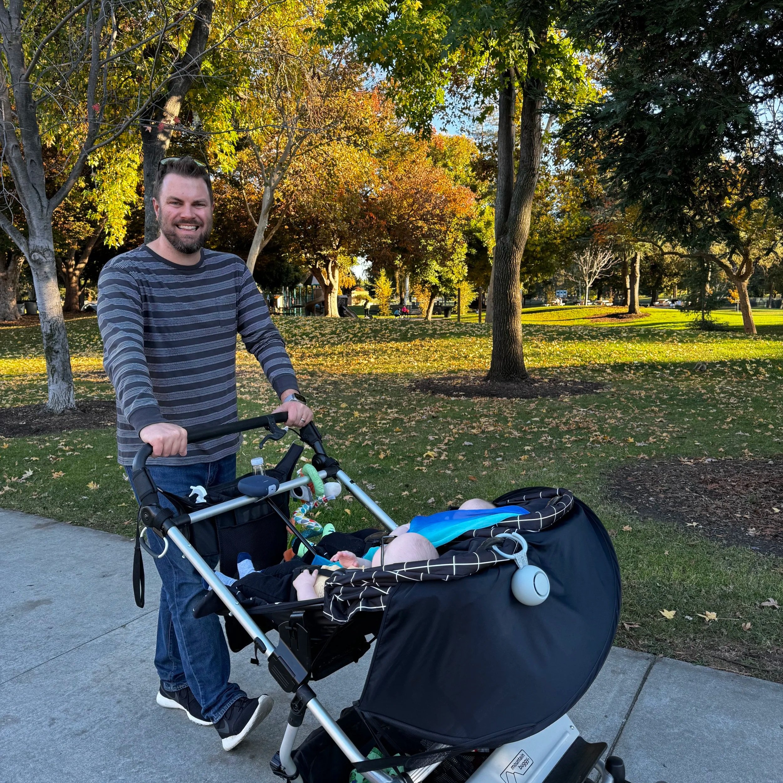A man with a beard and short hair smiling, standing outdoors in a park with trees that have autumn-colored leaves, pushing a stroller with a sleeping baby inside.