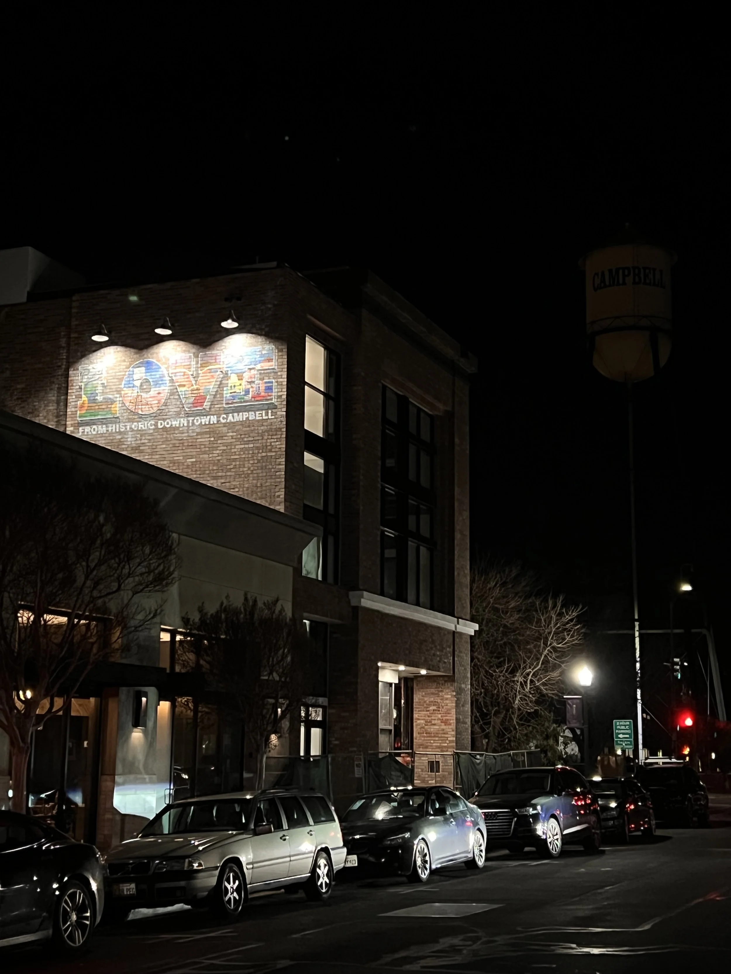 Night scene of a brick building with a mural reading 'LOVE FROM HISTORIC DOWNTOWN CAMPBELL' and a water tower with 'CAMPBELL' signage, several parked cars in front, and leafless trees.
