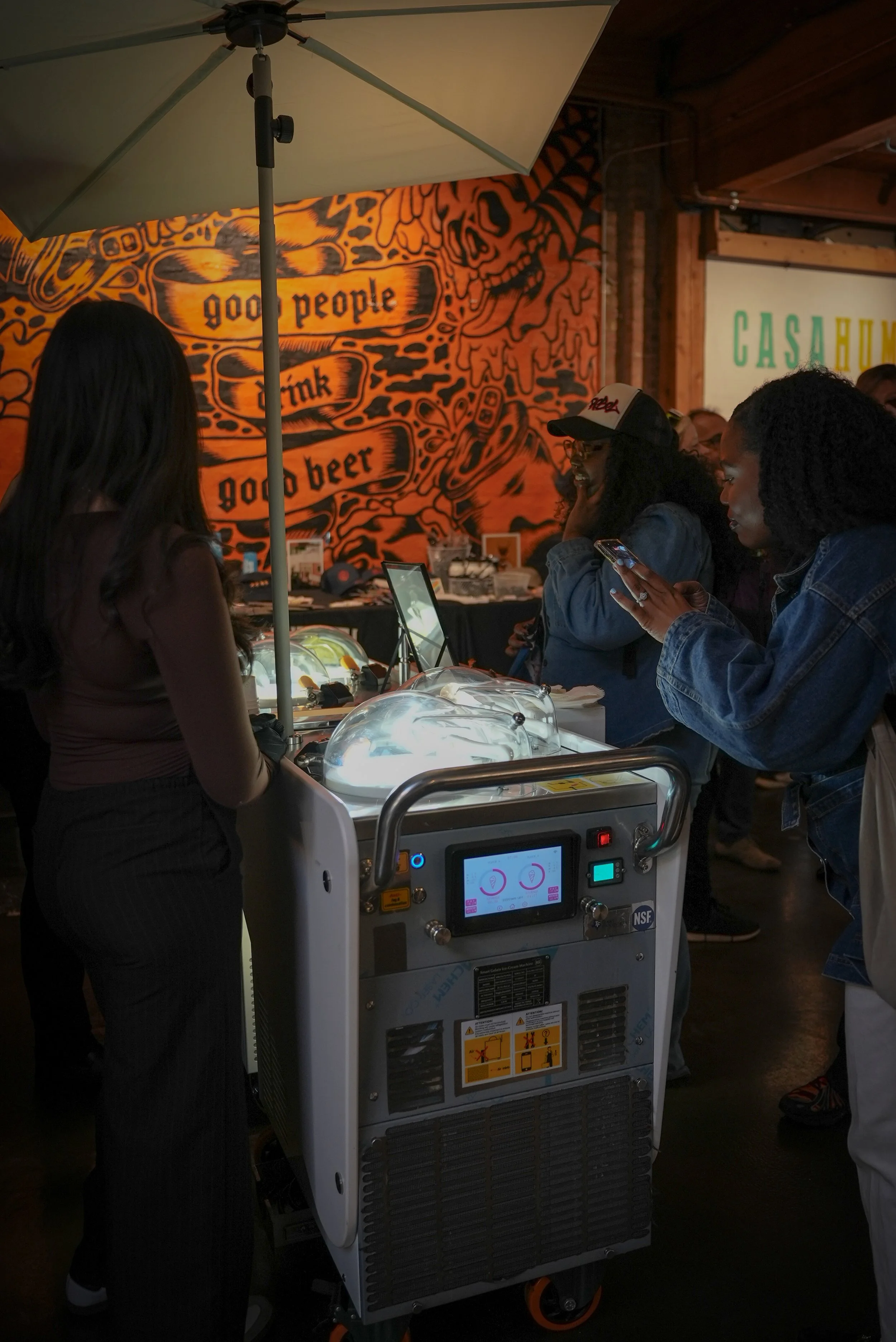 A group of people standing and interacting around an ice cream machine inside a restaurant or bar with orange and black wall art and a hammock-style sign in the background.