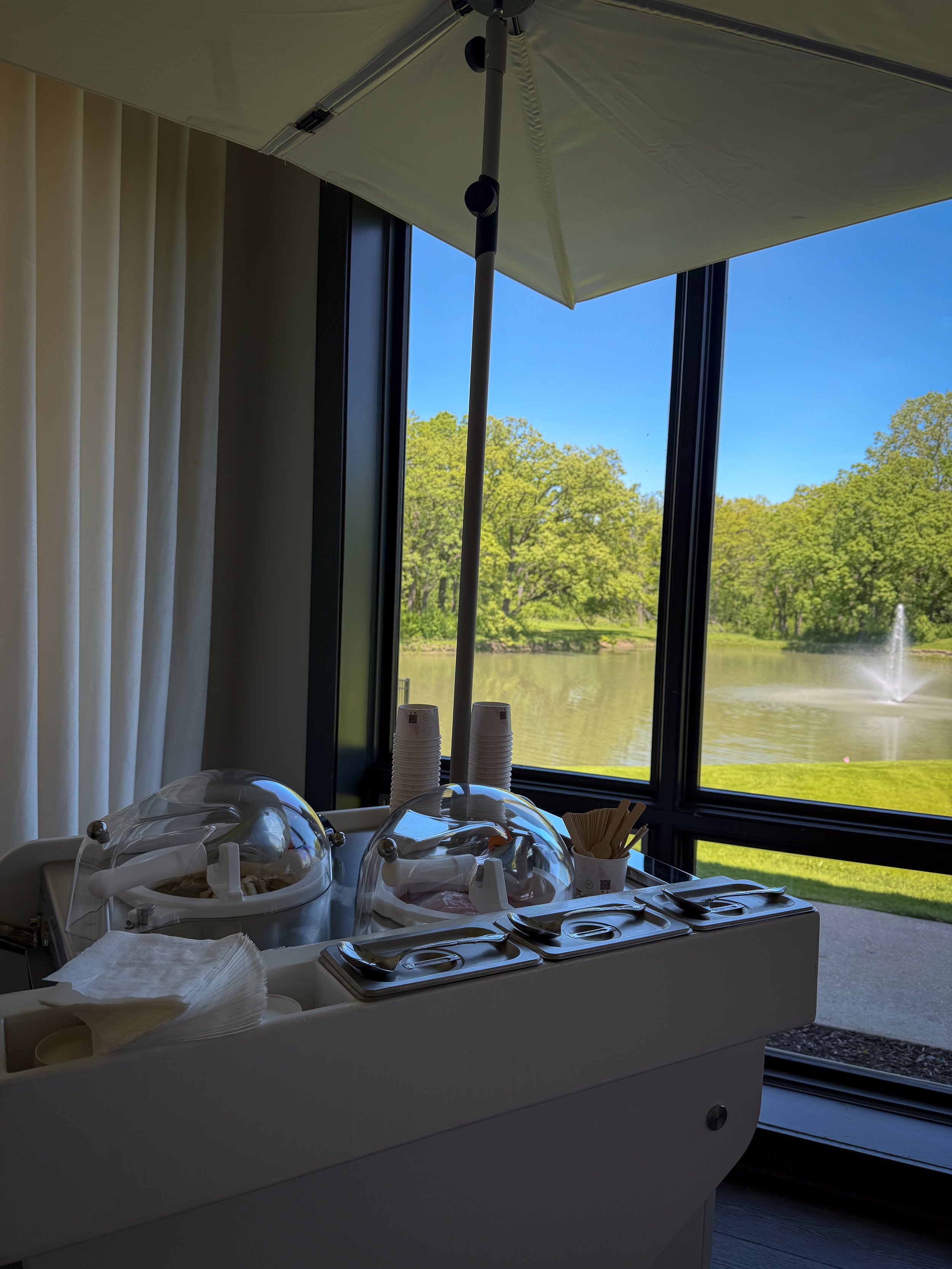 Ice cream buffet with toppings and cups near a large window showing trees and a lake on a sunny day.