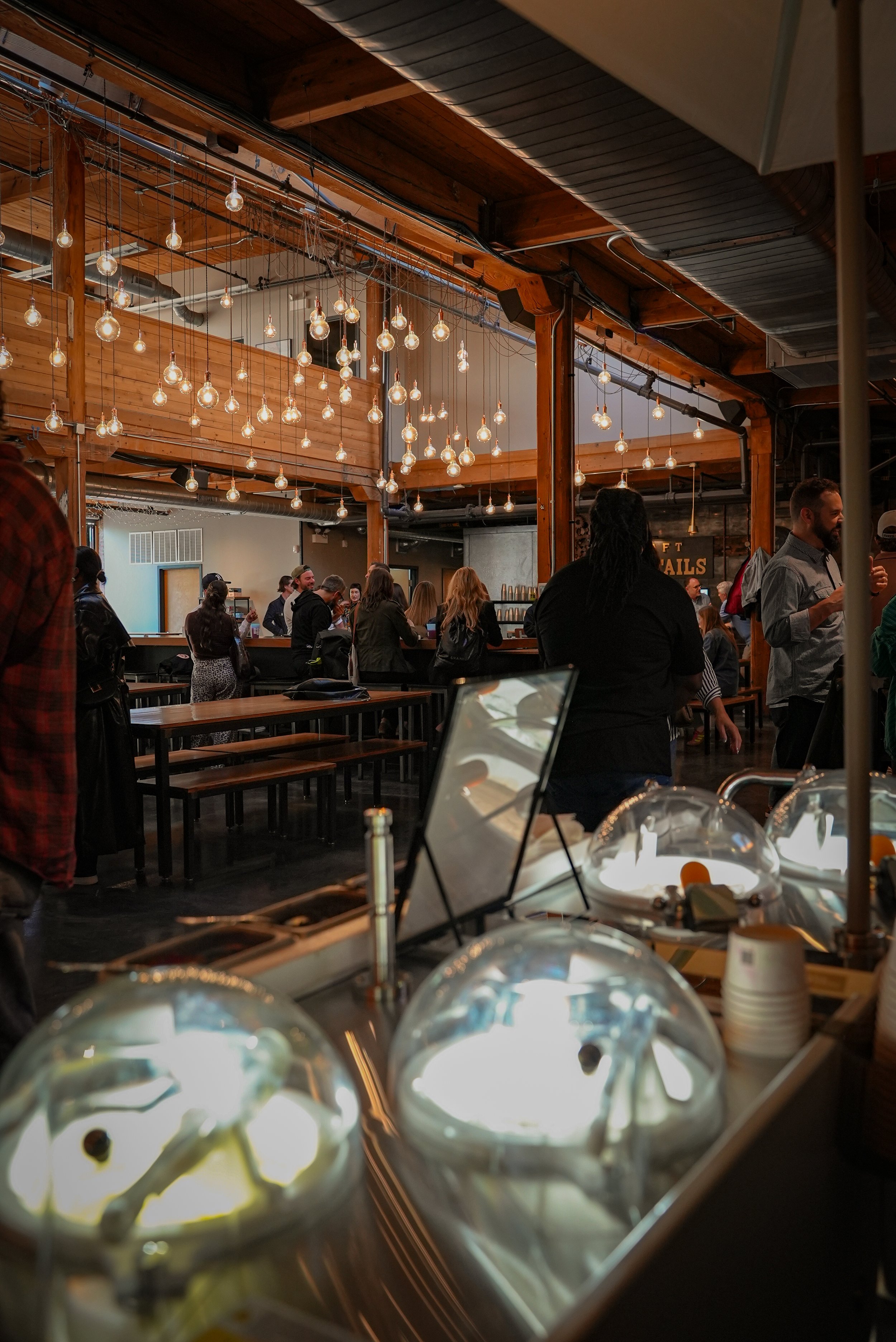 Inside a restaurant or bar with warm wooden decor and hanging Edison-style light bulbs, there are people standing and socializing, and there are condiments and desserts in the foreground.