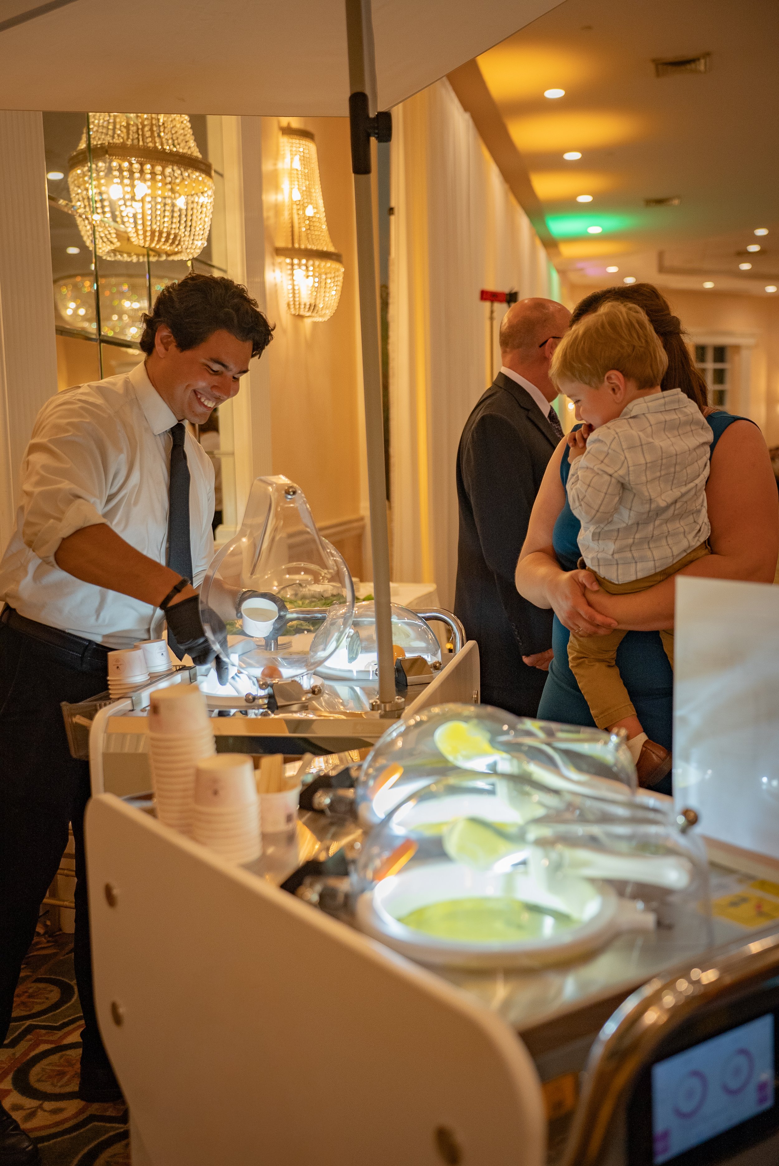 A waiter in a white shirt and black tie serving ice cream at a buffet table. In the background, a woman holding a young boy and an older man in formal attire are in a decorated, well-lit banquet hall.