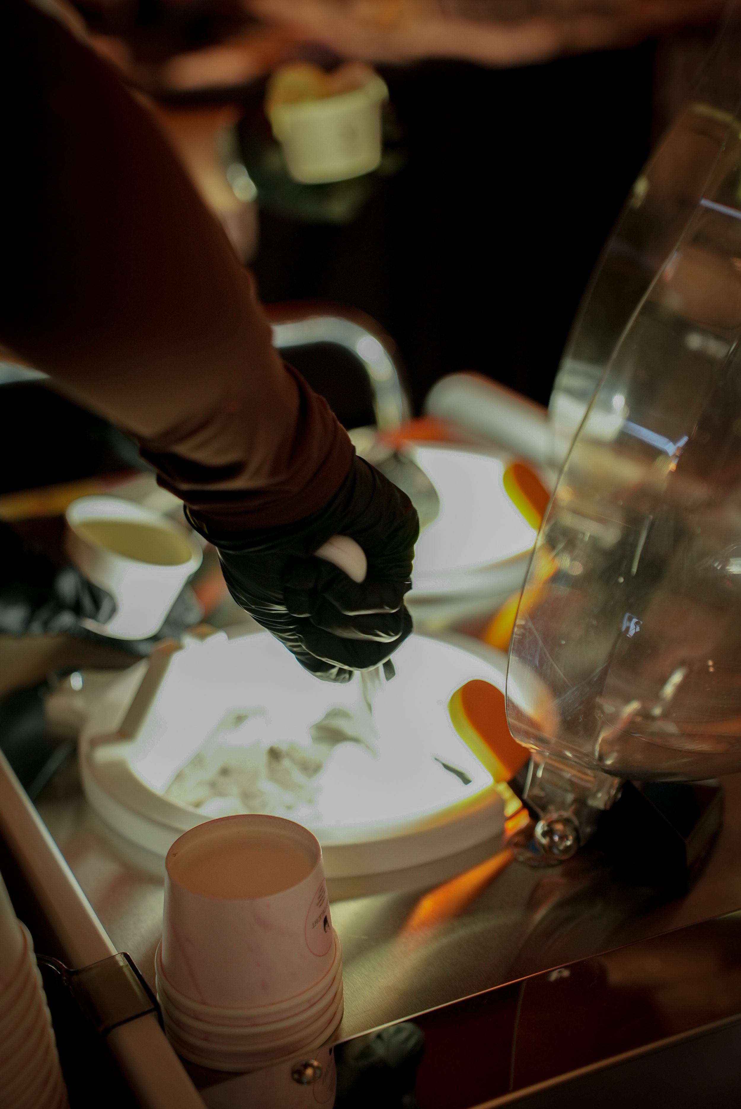 A person wearing black gloves using a spoon to serve ice cream from a machine in a setting that resembles a frozen yogurt shop.
