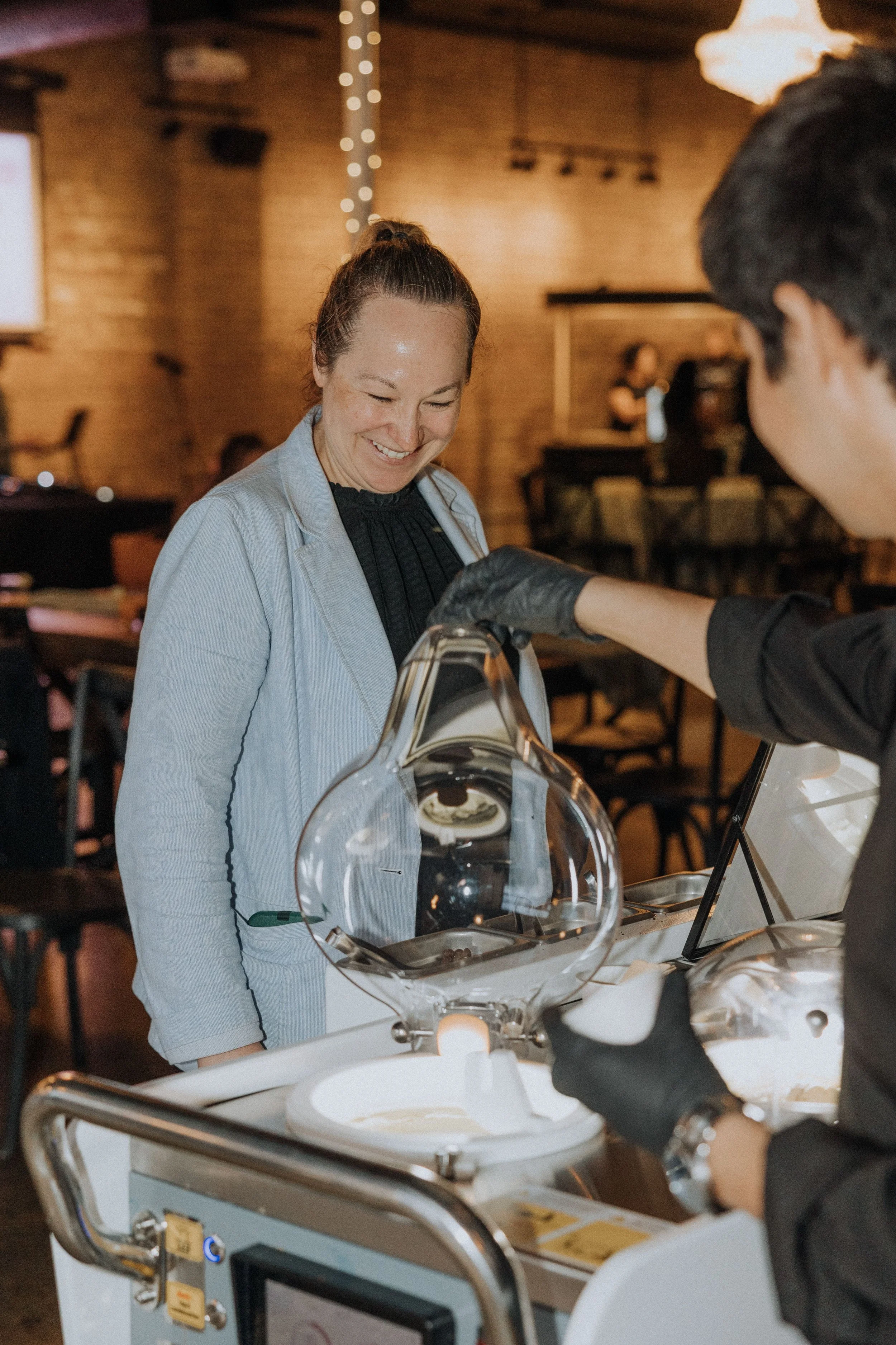 A woman smiling at a science exhibit with a glass and metal apparatus, while another person wearing black gloves appears to be explaining or demonstrating something to her in a dimly lit indoor space with wooden walls.