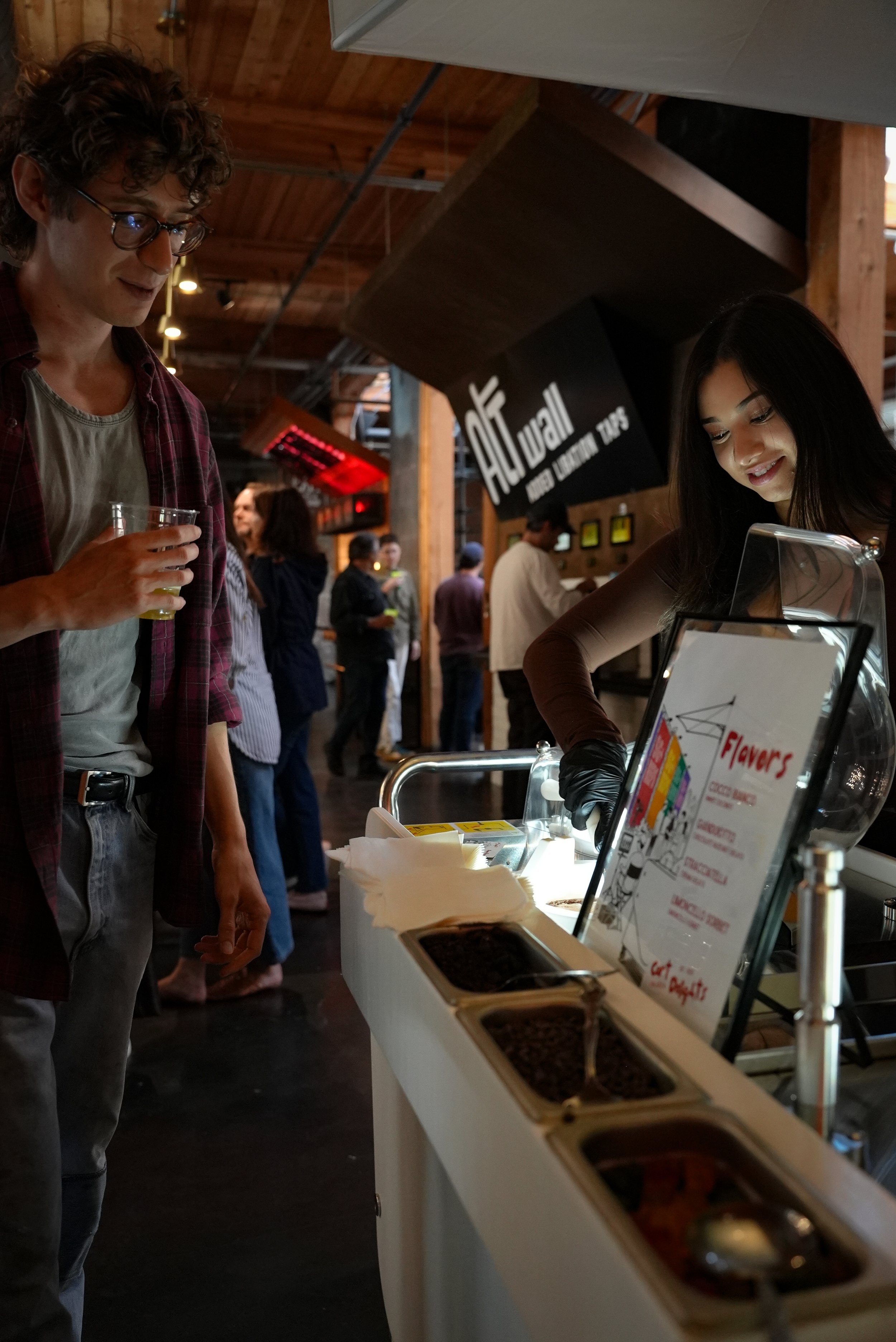 A man holding a drink and a woman serving toppings at a dessert or ice cream stand inside a wooden venue with people in the background.