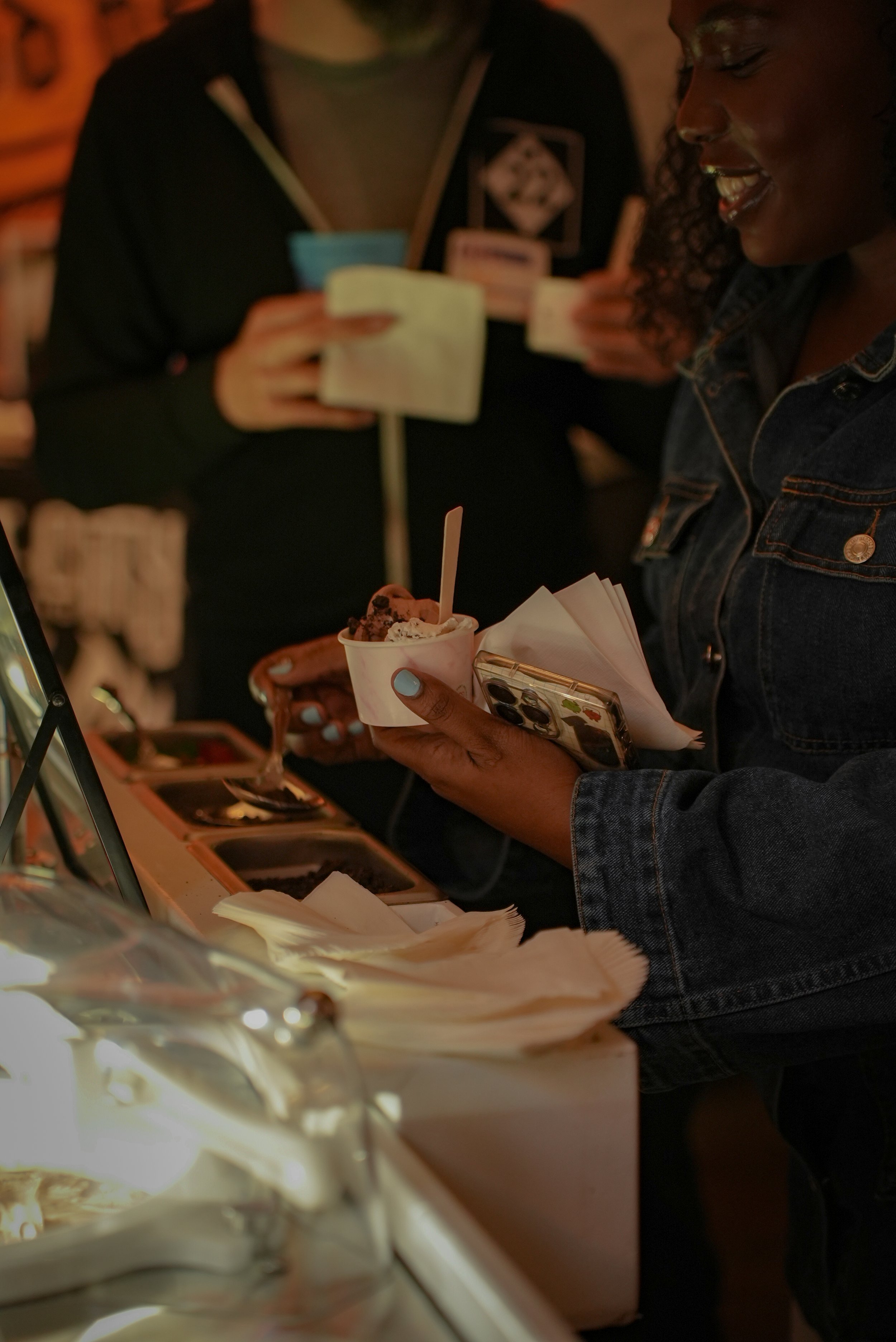 People at an ice cream shop selecting flavors, with one person holding a cup of ice cream and a phone, and others carrying cups or containers.