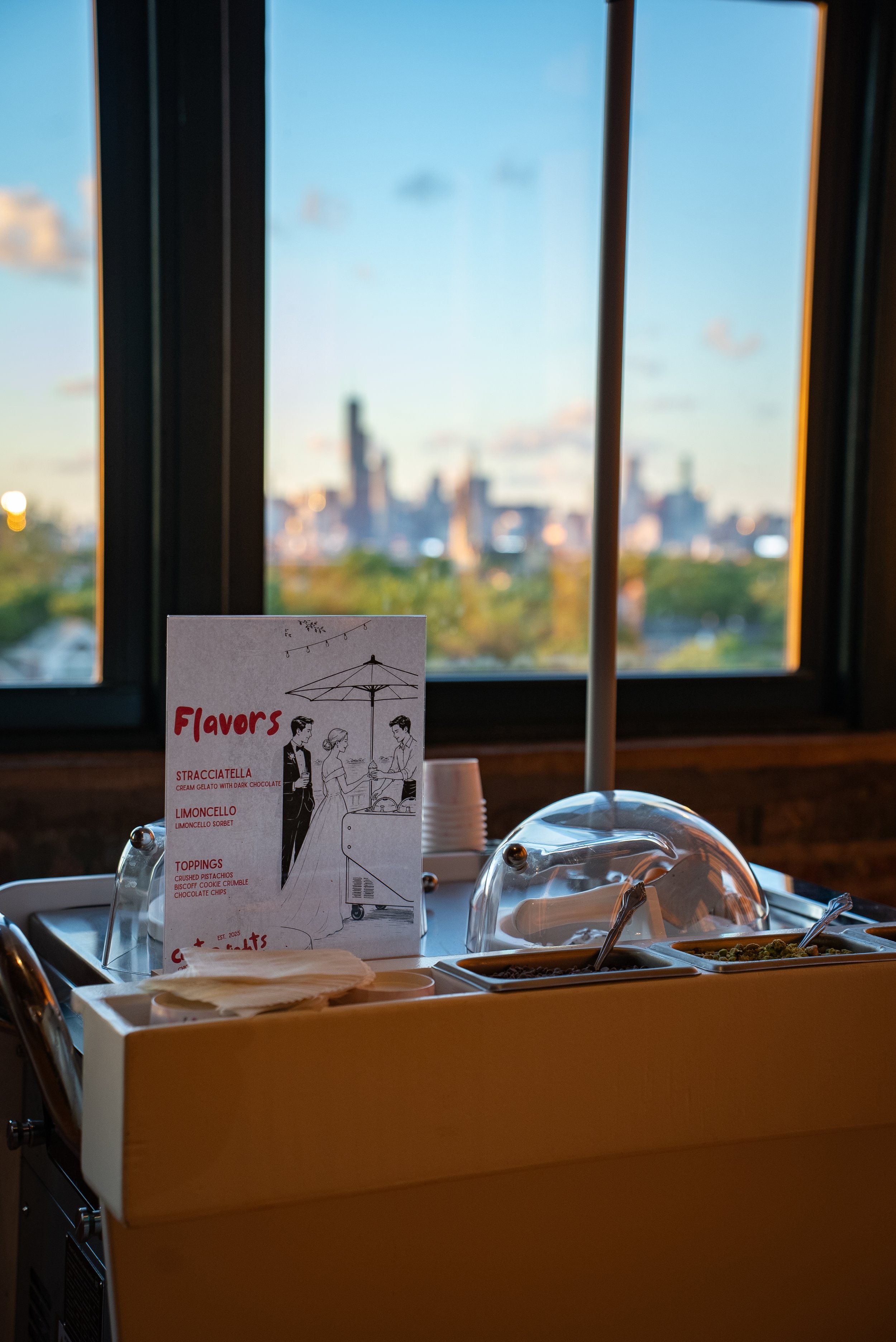 A buffet table with various dishes and a menu card in front, with a large window in the background showing a city skyline at sunset.