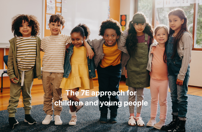 A group of children stand together in a class room setting arms over one anothers shoulders smiling at the camera
