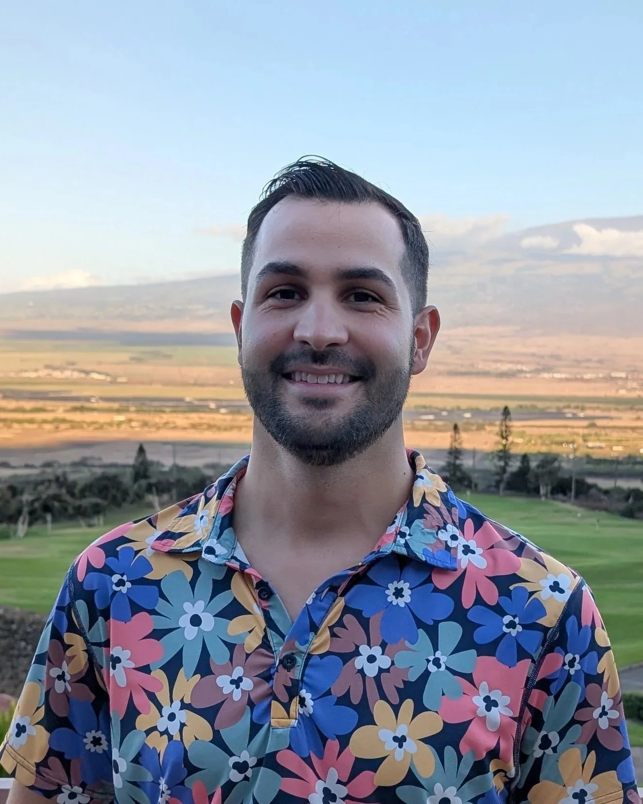 A man standing outdoors in front of a landscape with fields, hills, and a blue sky, wearing a colorful Hawaiian shirt with pink, blue, yellow, and white flowers.