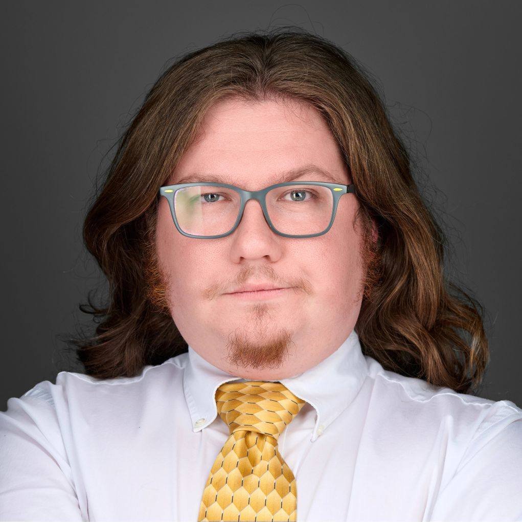 A man with long brown hair, glasses, a goatee, and a serious expression. He is wearing a white shirt and a yellow tie with a honeycomb pattern against a gray background.