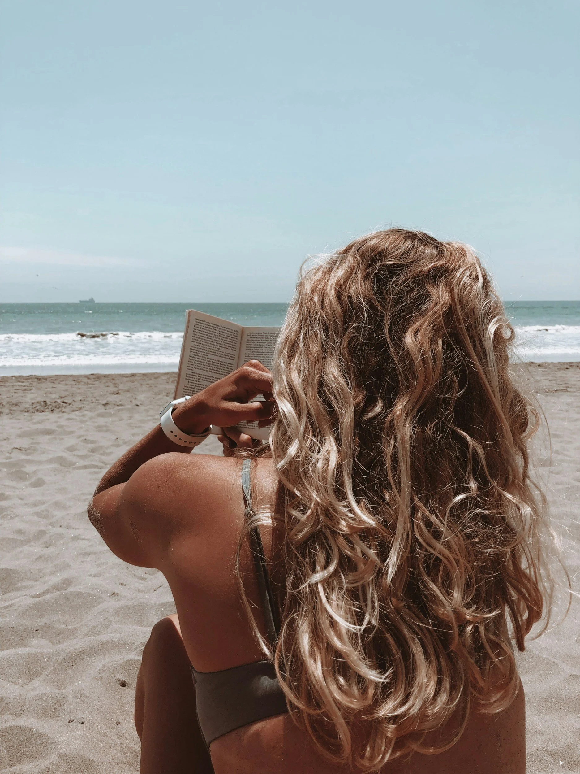 A woman with long, curly blonde hair is reading a book while lying on the beach. The ocean waves and a ship are visible in the background.