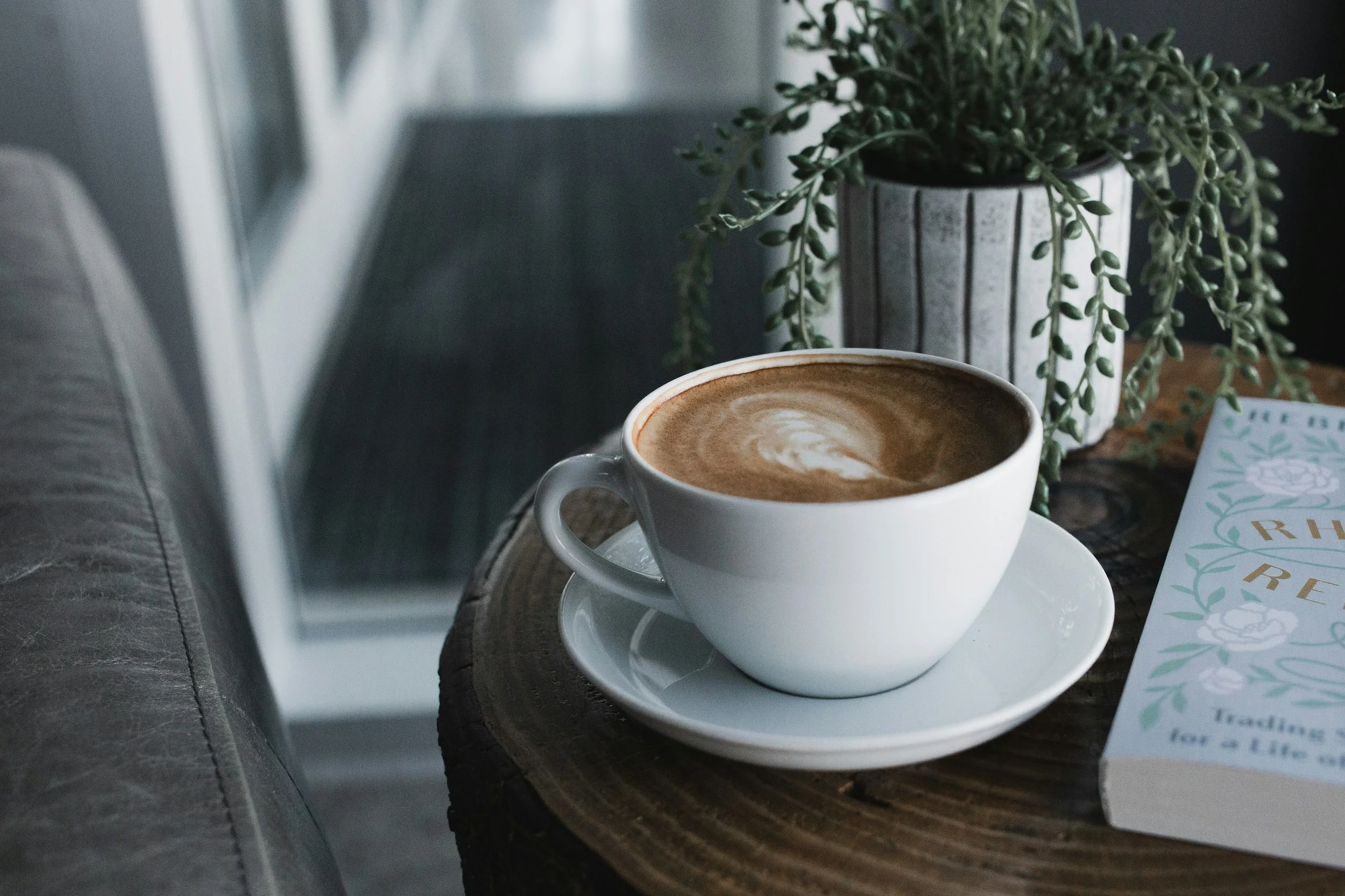 A white coffee cup filled with a frothy latte sits on a white saucer on a wooden table, with a potted green plant and a magazine nearby.