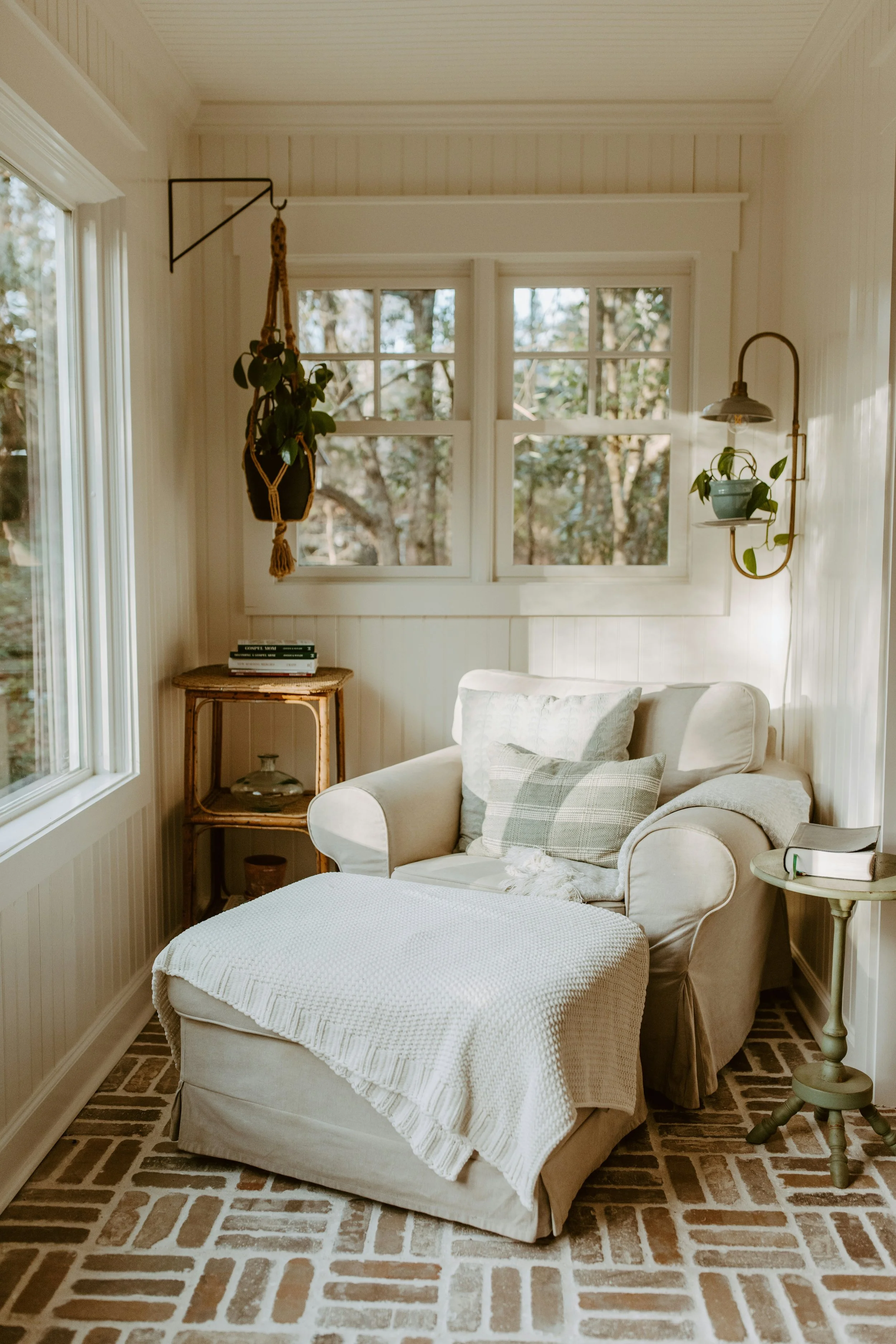 Sunlit cozy living room with white walls, a beige armchair with a white knitted throw and pillows, a small side table, hanging plants, and a brick floor.