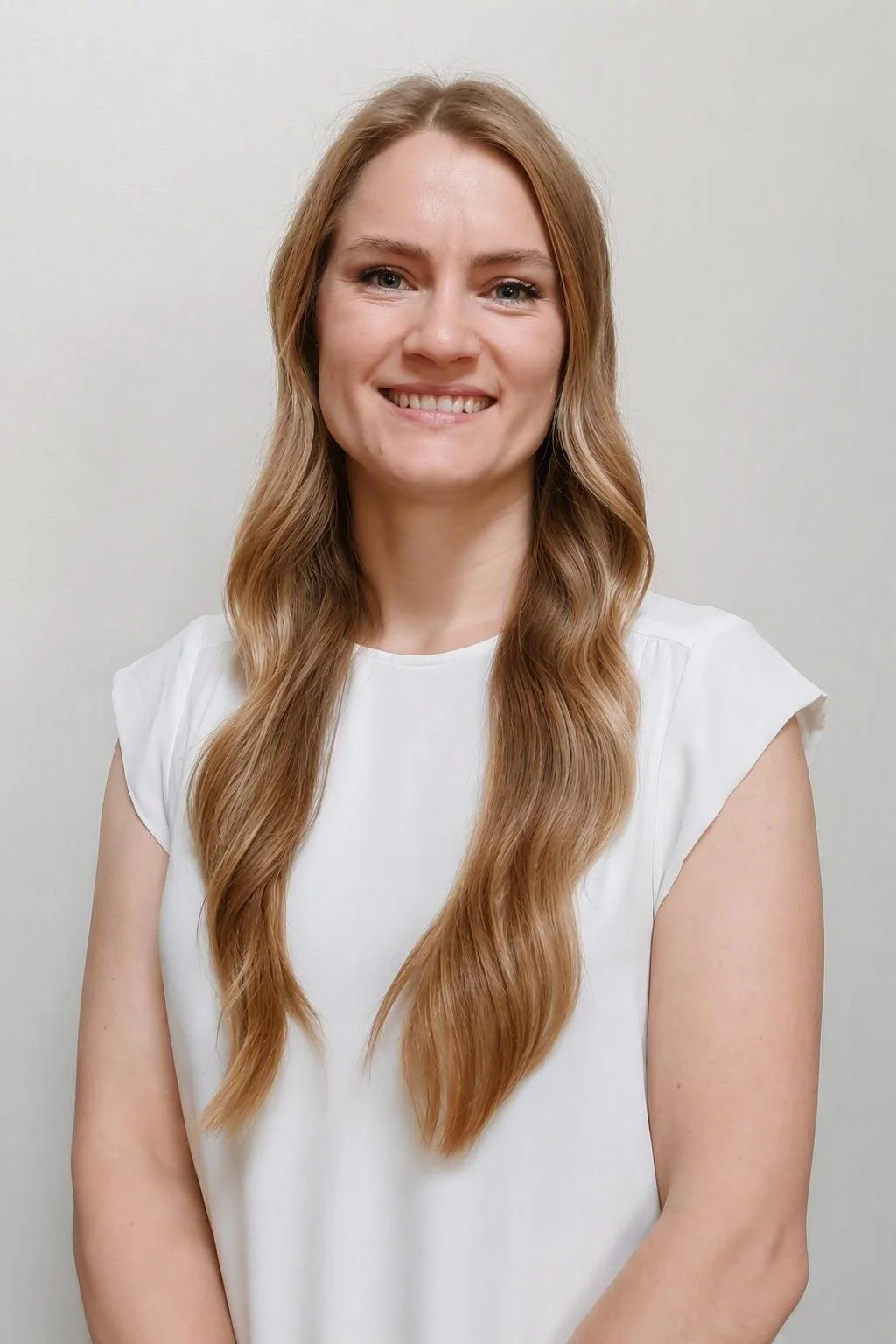 Portrait of a young woman with long wavy blonde hair, smiling, wearing a white sleeveless top, standing against a plain light-colored background.