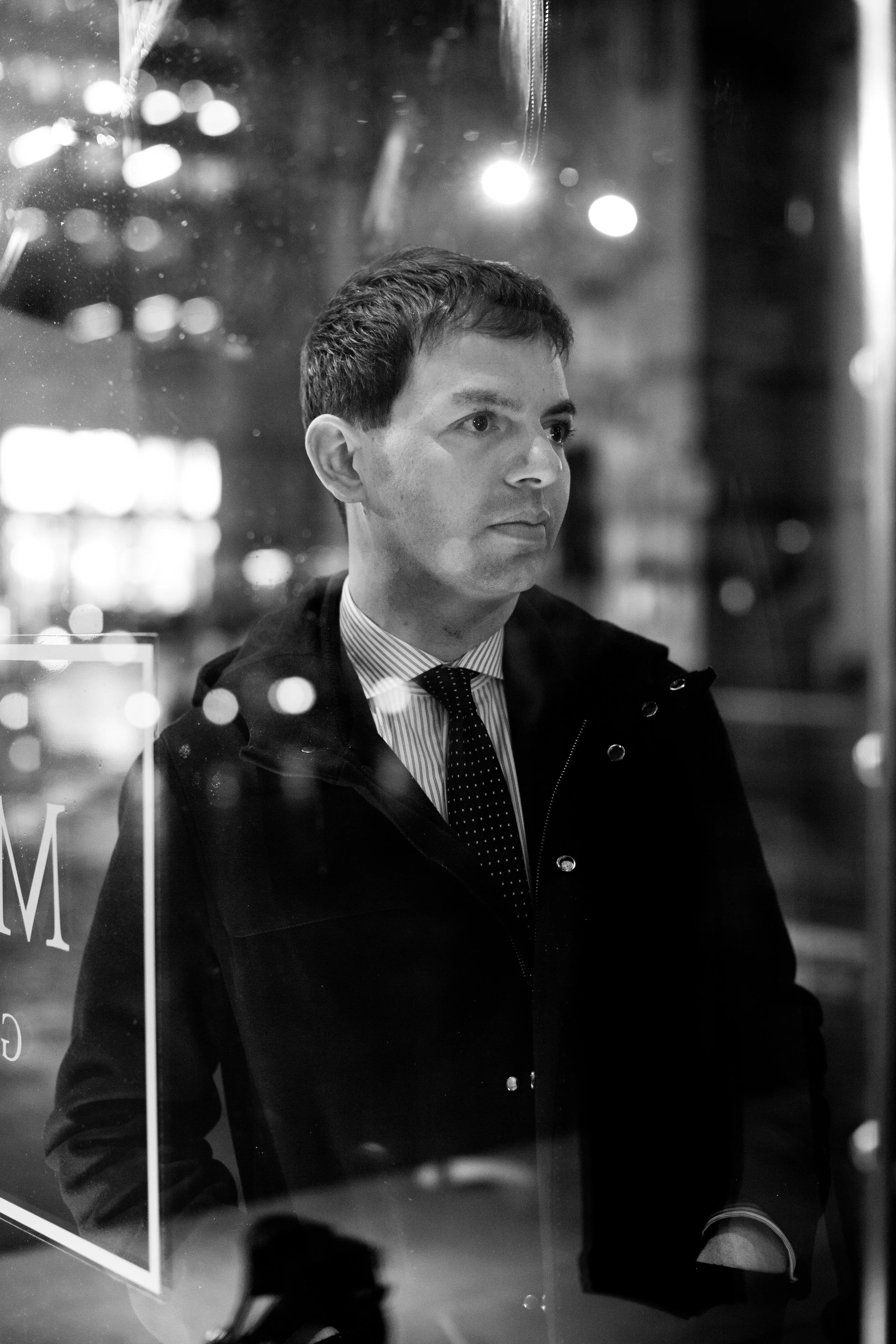 Dan Nimmer dressed in a collared shirt, tie, and dark jacket, stands outside near a glass window with reflections of bokeh lights in the background.