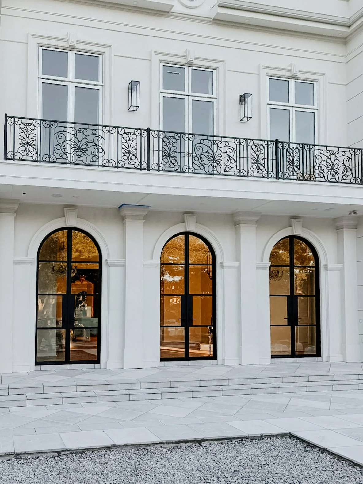 A white multi-story building with large arched windows and a decorative black wrought iron balcony on the upper floor. The windows on the lower level are tall with black frames, and there are steps leading up to the entrances.