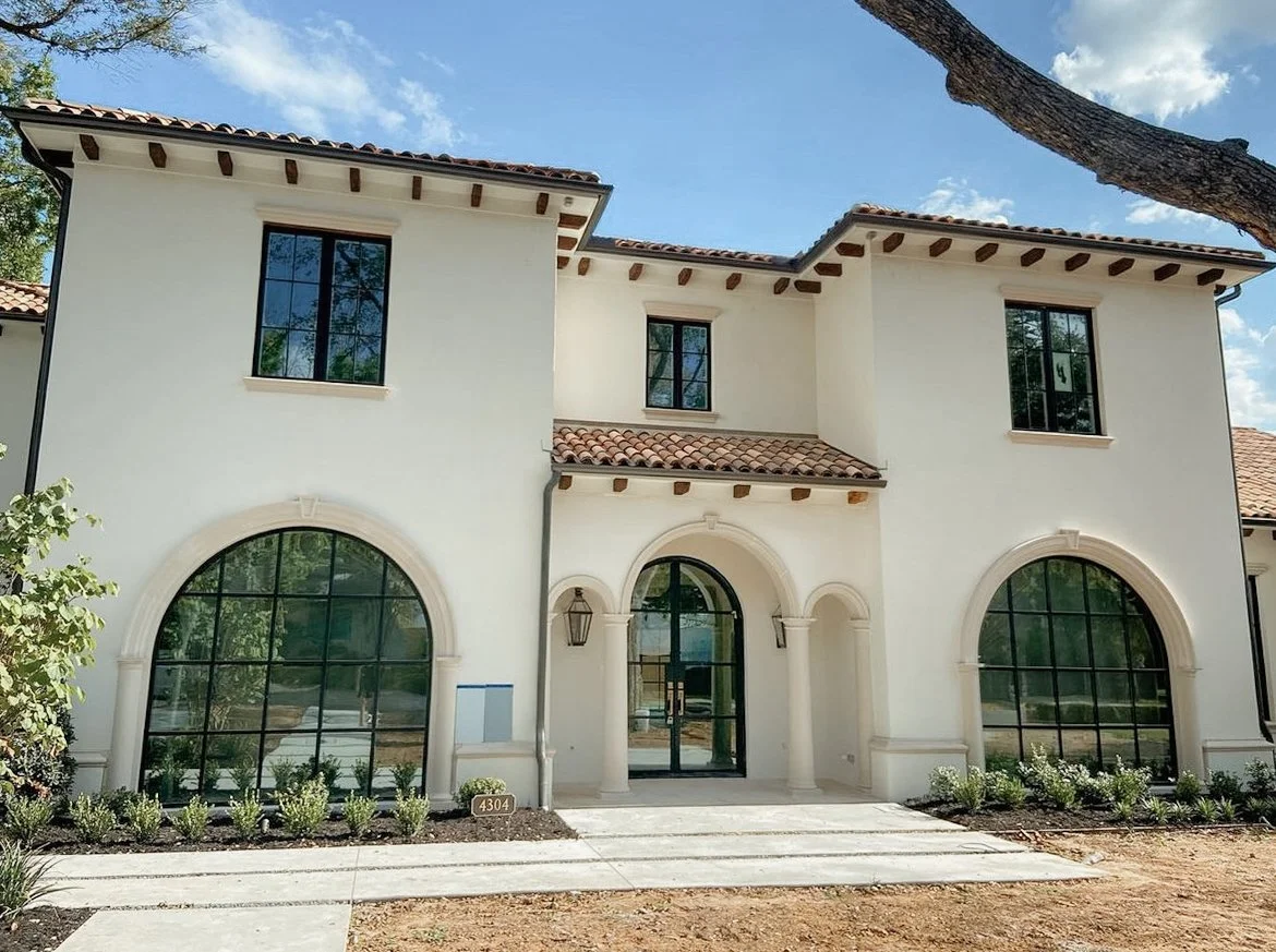 Modern two-story house with white stucco walls, arched windows, and a tiled roof, surrounded by landscaping.