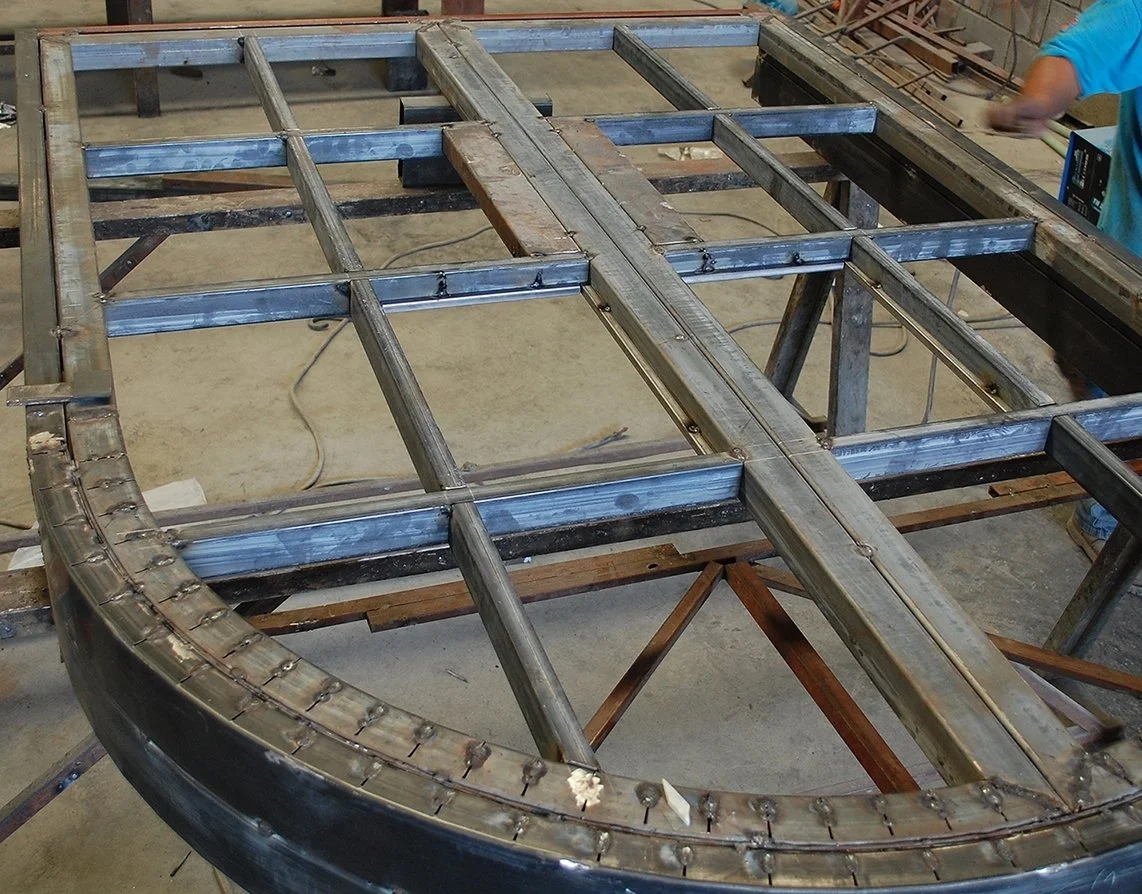 Metal framework structure under construction, with a curved metal piece in the foreground, and a person working on the right side.