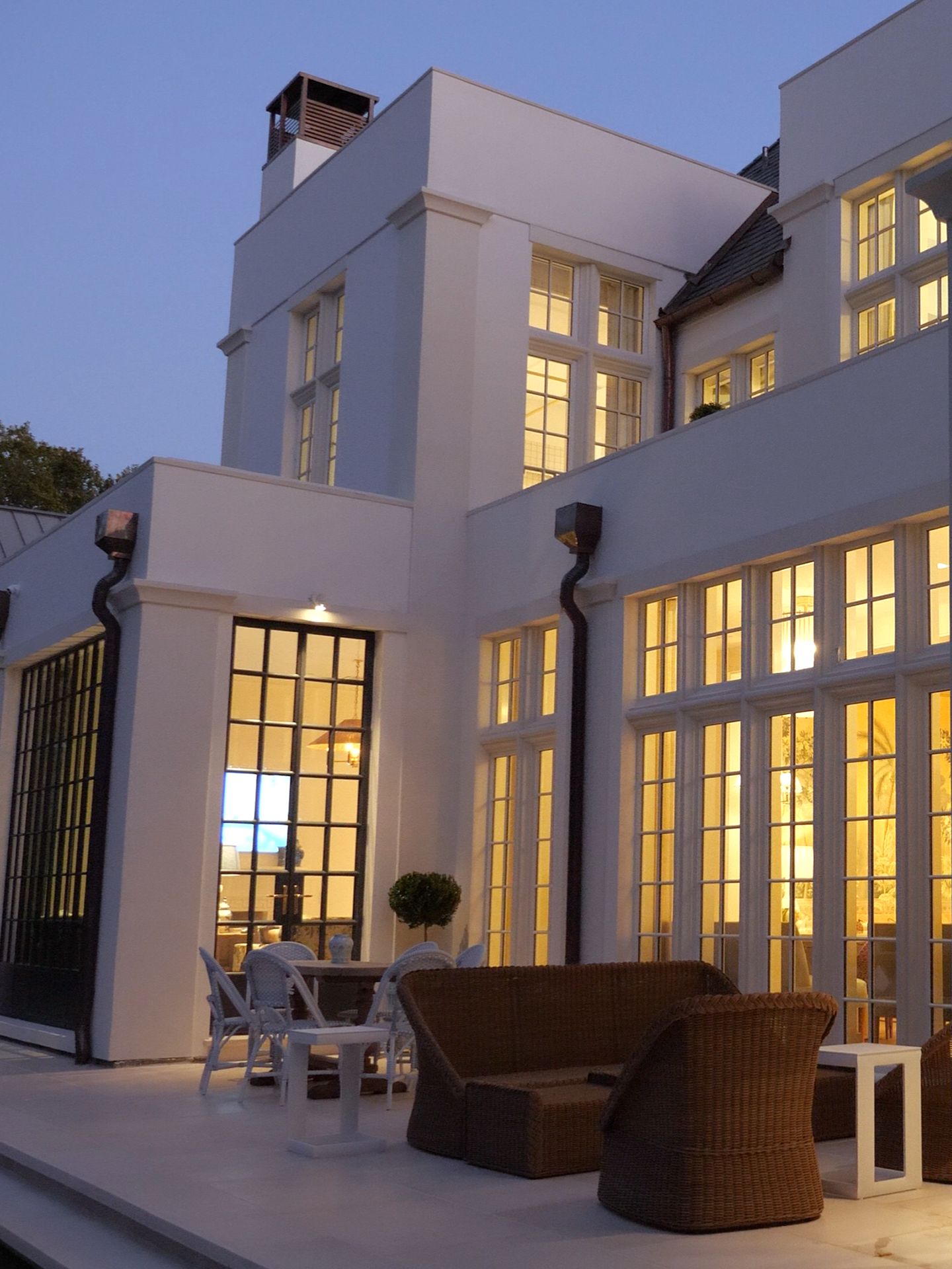 Modern house with large glass windows and outdoor patio furniture, illuminated interior, evening setting.