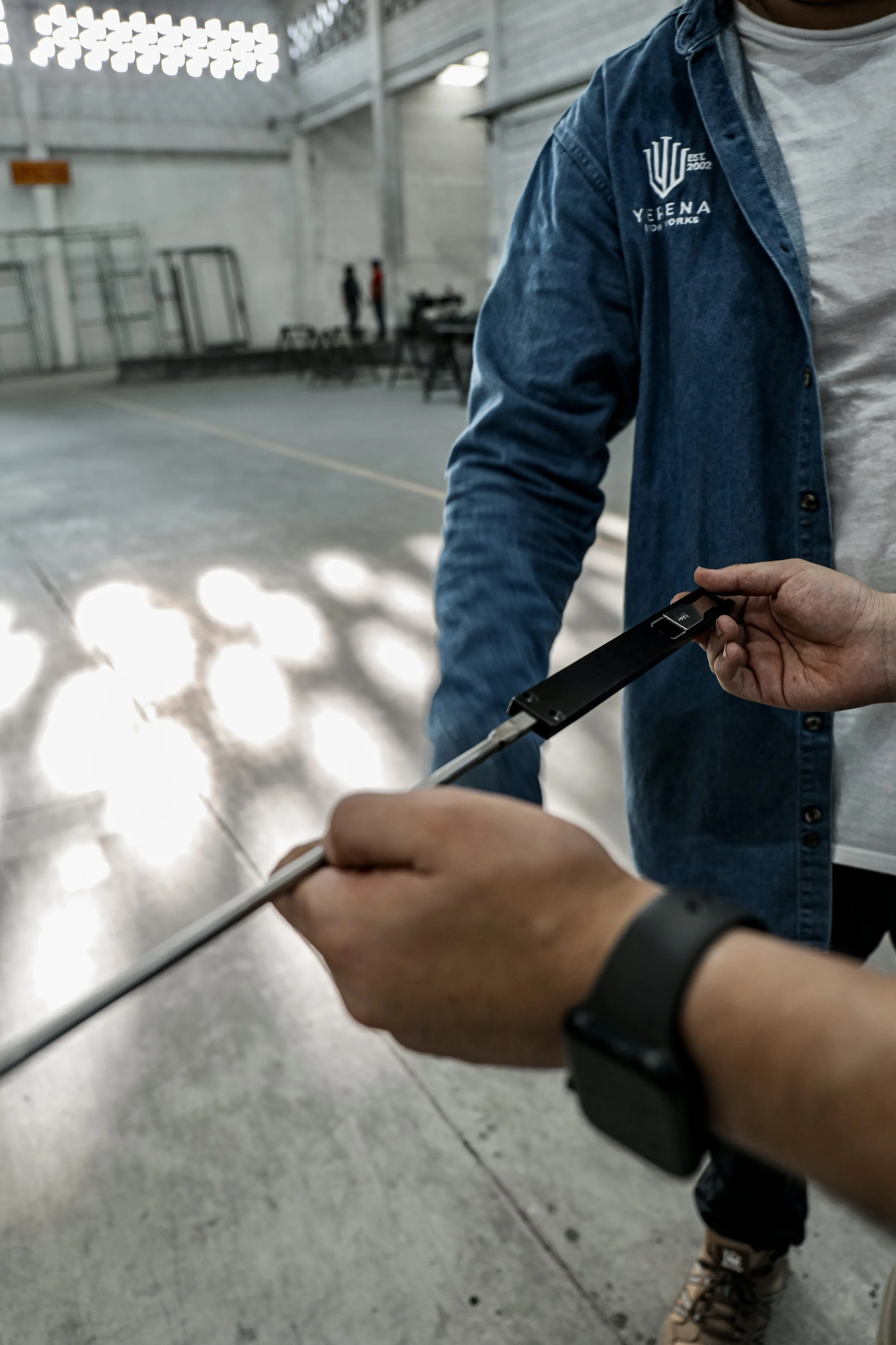 Close-up of two people exchanging a black key fob, with one person wearing a smartwatch and the other wearing a denim jacket. The background shows a large indoor space with concrete floors and a couple of people in the distance.