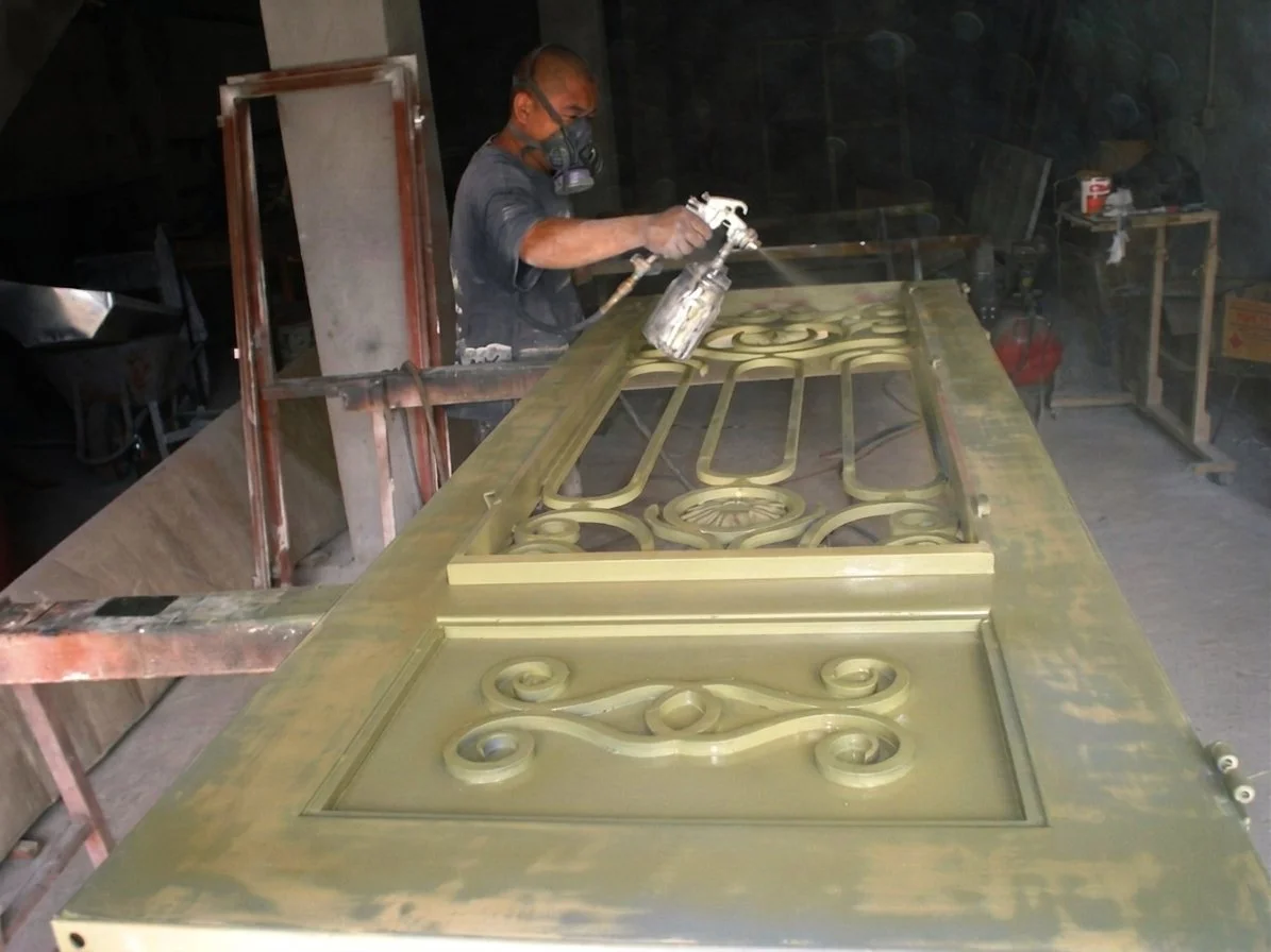A worker wearing a mask spray paints a large decorative metal door with ornate scrollwork.