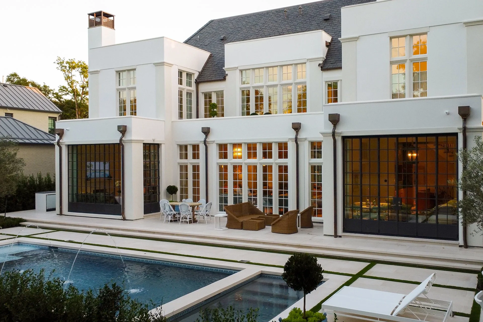 A modern white multi-story house with large glass windows, a patio with outdoor furniture, and a swimming pool with fountains, surrounded by greenery.