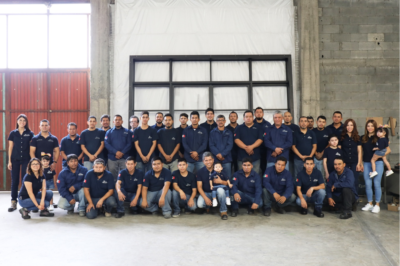 A large group of people wearing matching navy blue shirts and jackets, posing for a group photo inside a spacious industrial or warehouse space with concrete walls.