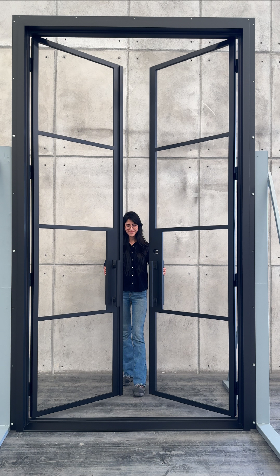 A woman standing behind a large, open black metal door frame, in front of a plain concrete wall.