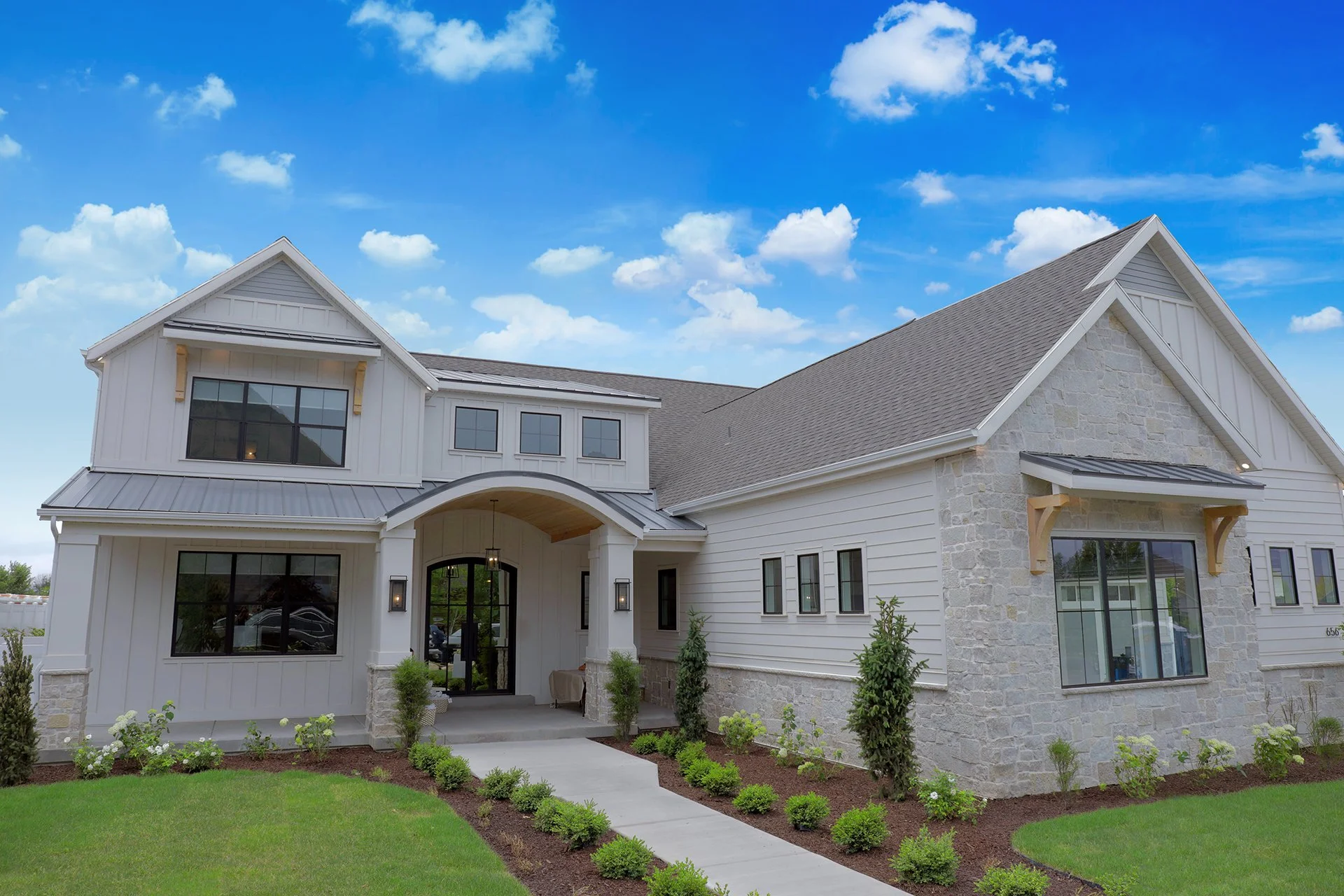 Front view of a modern white house with stone accents, large windows, and a gray shingle roof, surrounded by a neatly landscaped yard with a curved walkway and green grass.