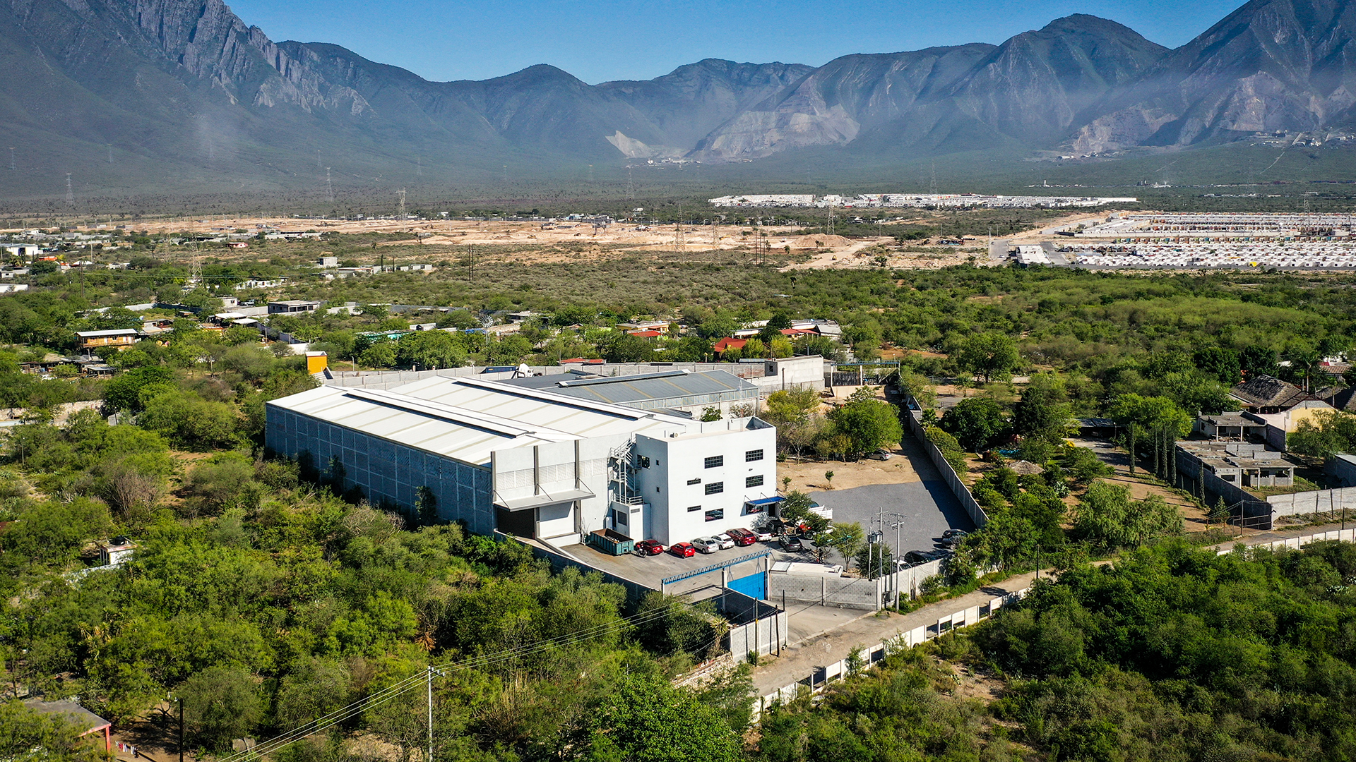 A wide landscape view showing a small town with a large industrial or commercial building surrounded by trees, with a mountain range in the background under a clear blue sky.