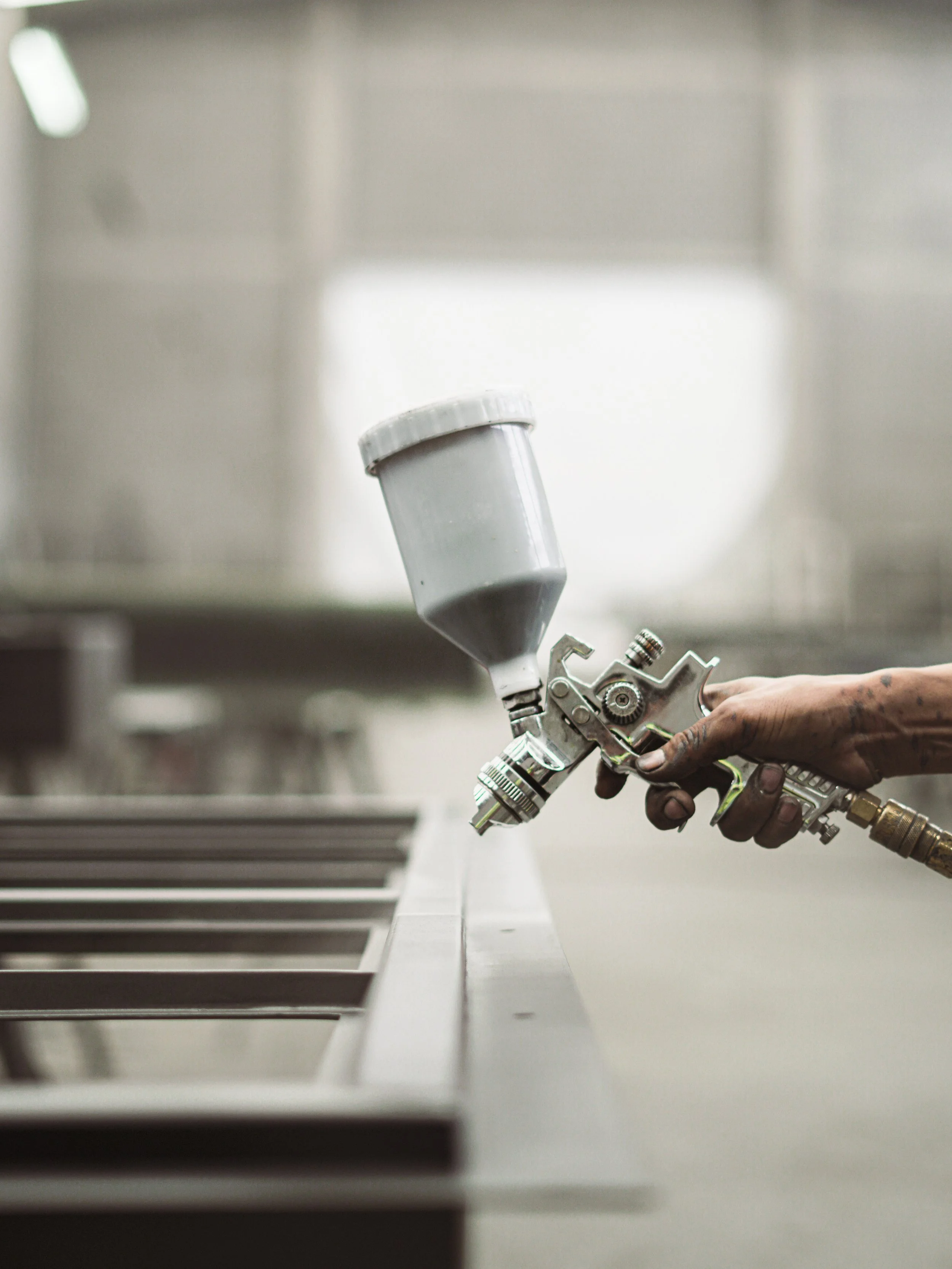 Close-up of a person's hand holding a spray gun, painting a white surface in an industrial setting.