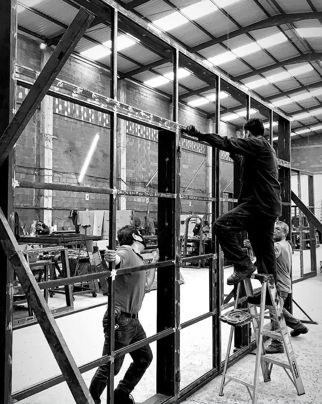 Three workers assembling a metal frame structure inside a large industrial warehouse.