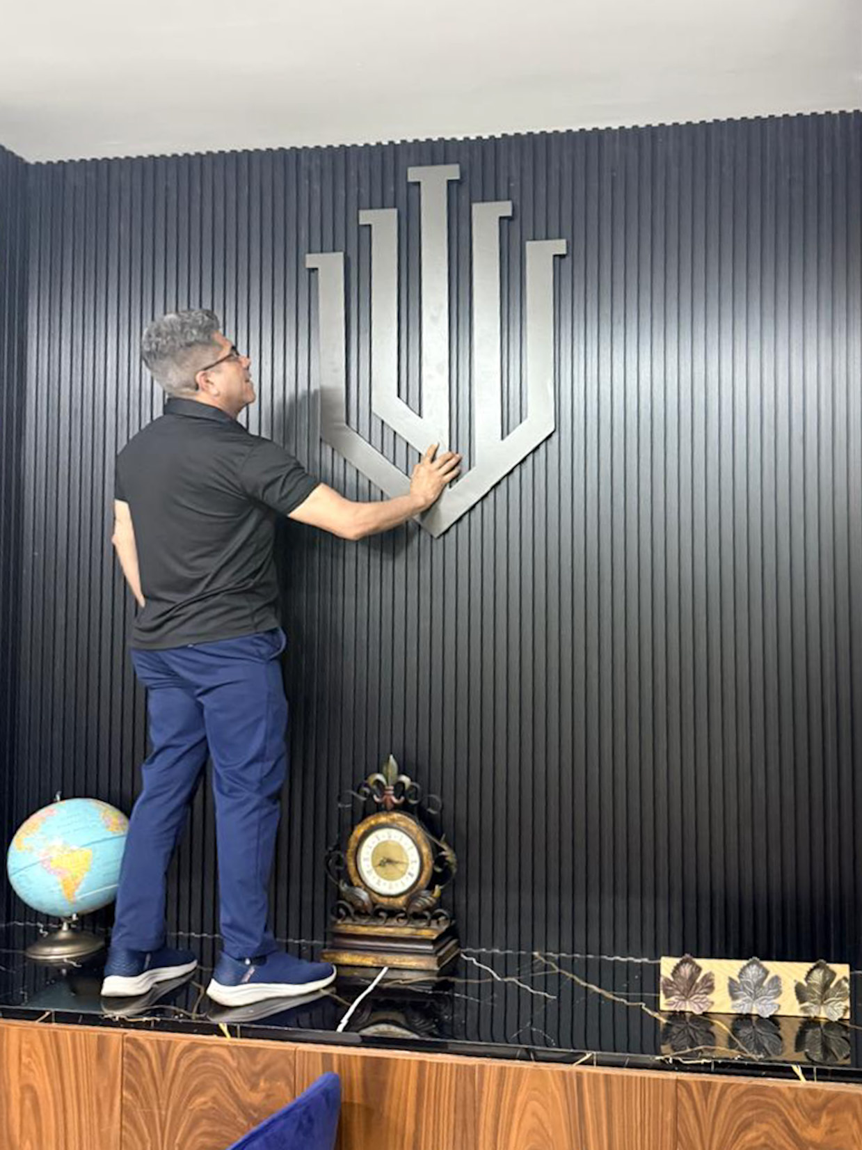 A man adjusting a large silver Indiana University logo on a dark, vertical-slatted wall. Below, there is a globe, an antique clock, and a decorative leaf pattern on a wooden surface.