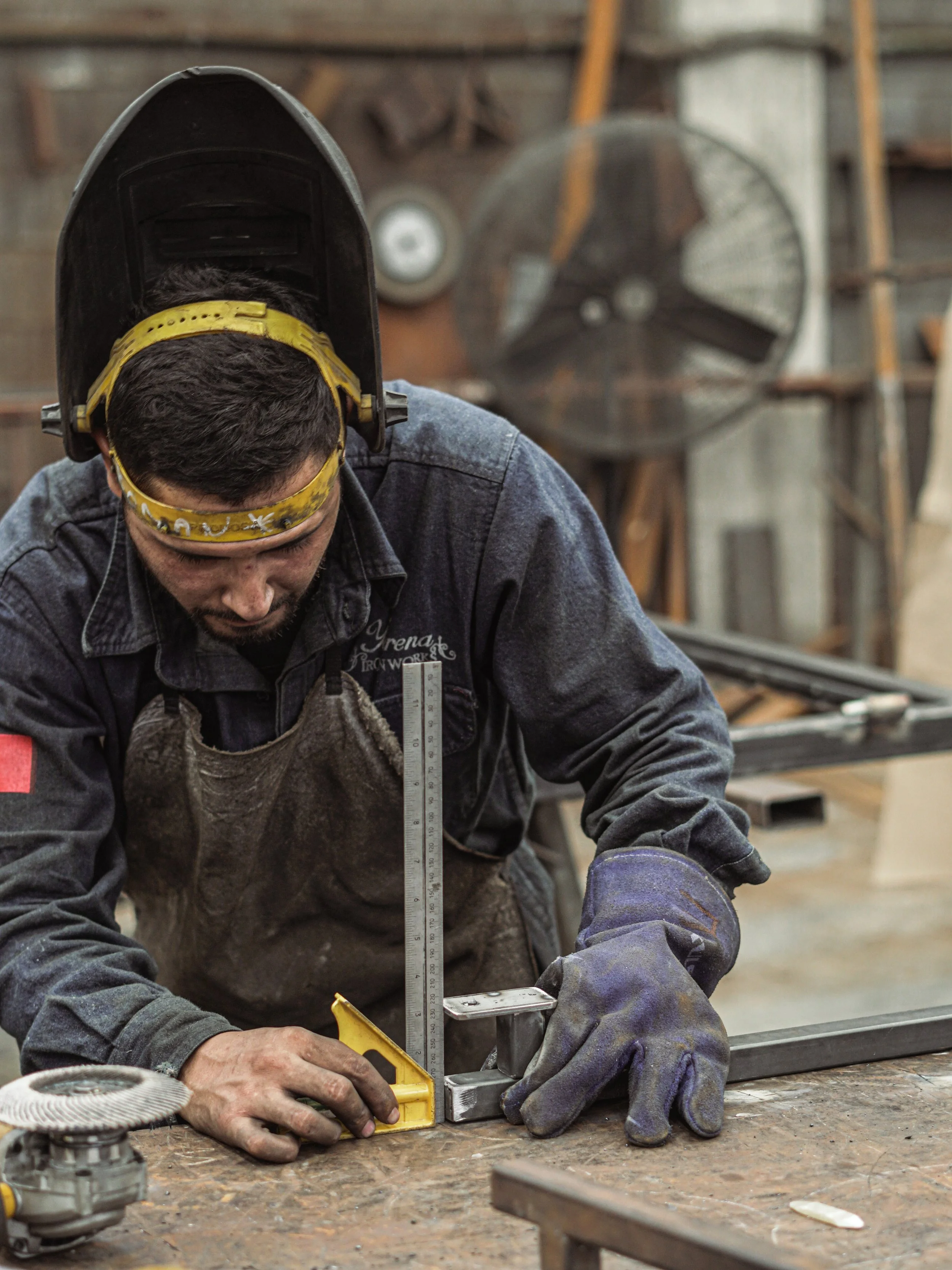 A man wearing a welding helmet and gloves working with metal parts in a workshop, using a measuring tool.