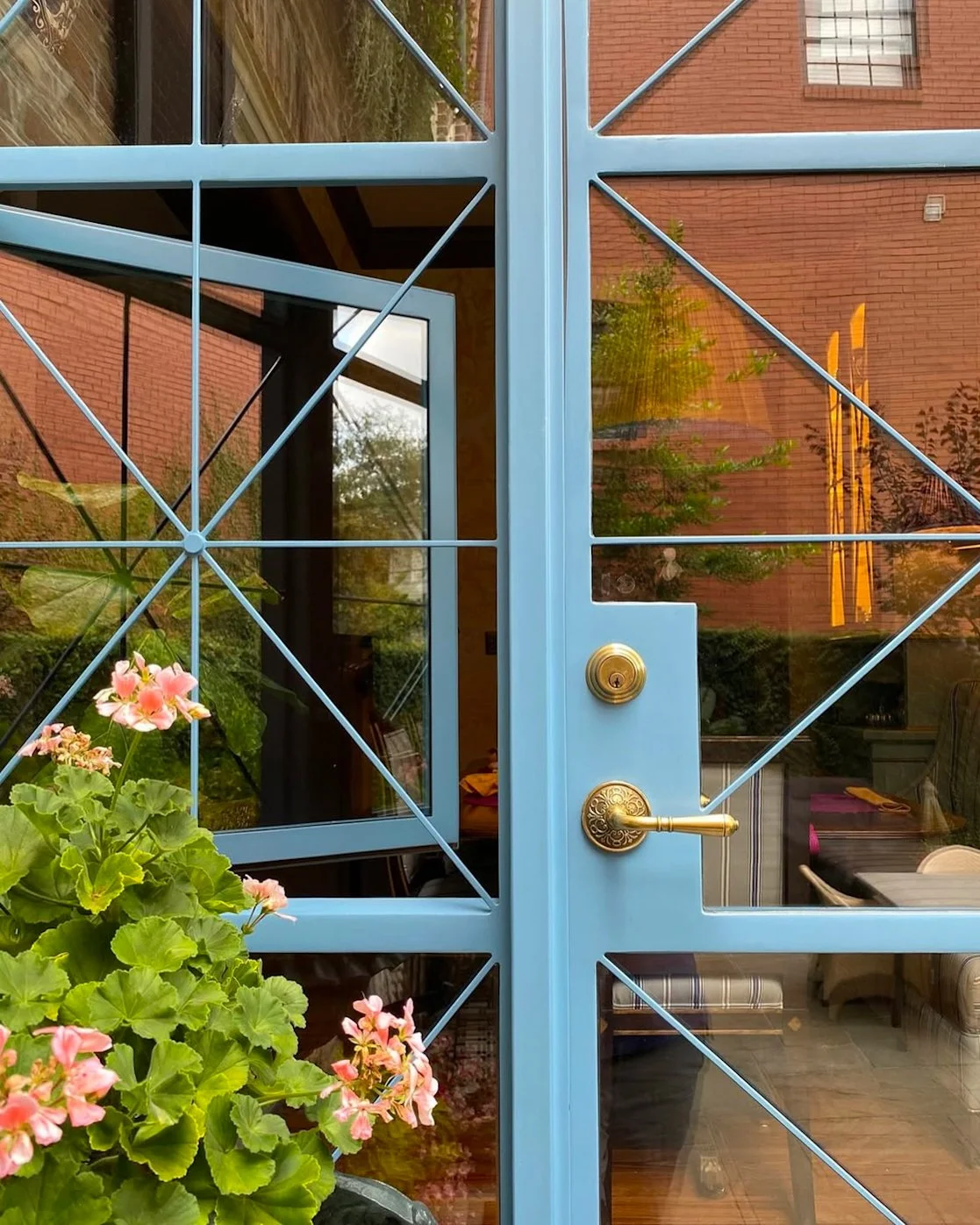 Blue front door with decorative lattice design, gold handle and lock, glass panels revealing a dining area with chairs and a table inside, and pink flowers in a pot outside.