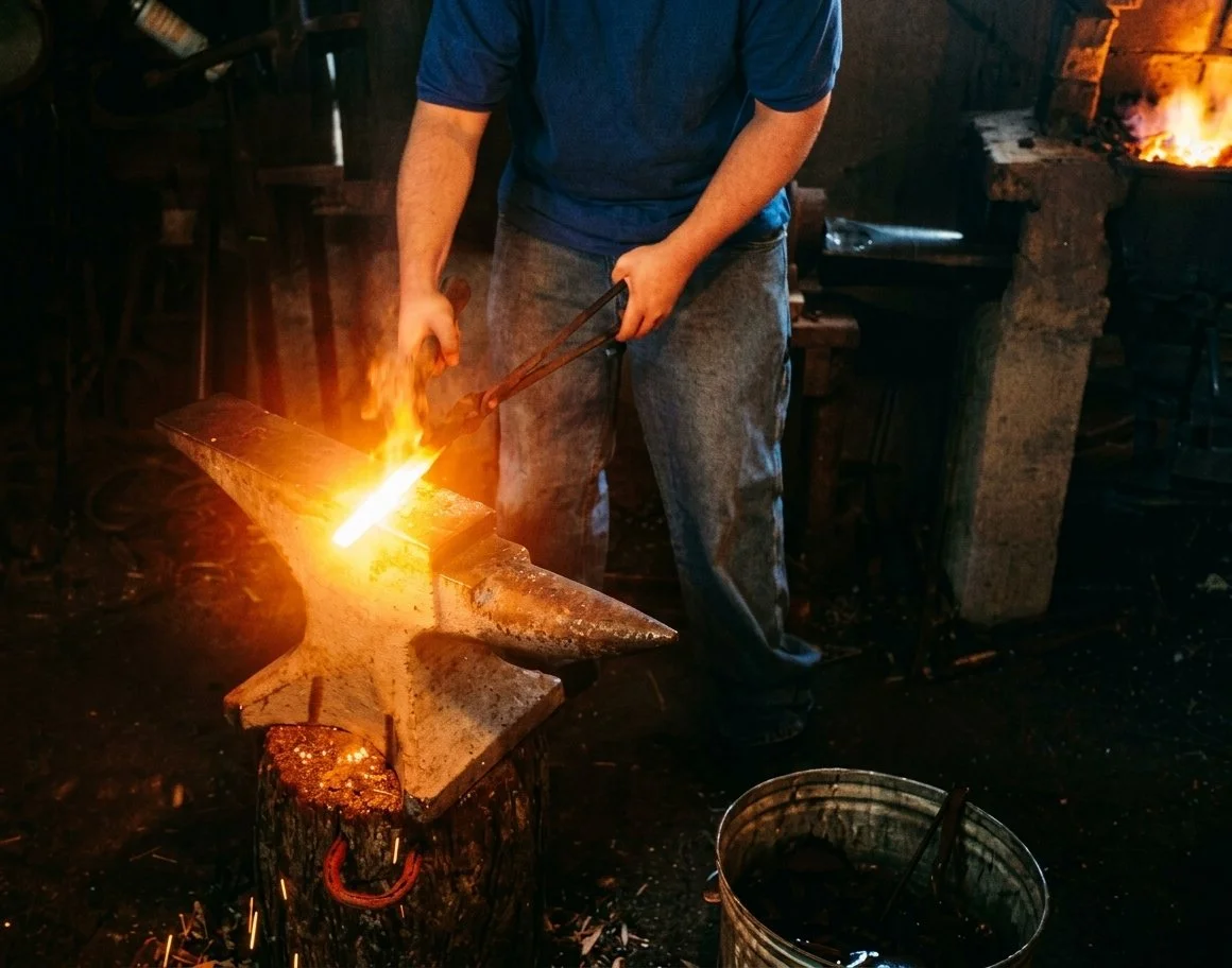 A blacksmith in a workshop heating metal on an anvil with a forge flame, wearing a blue shirt and jeans.