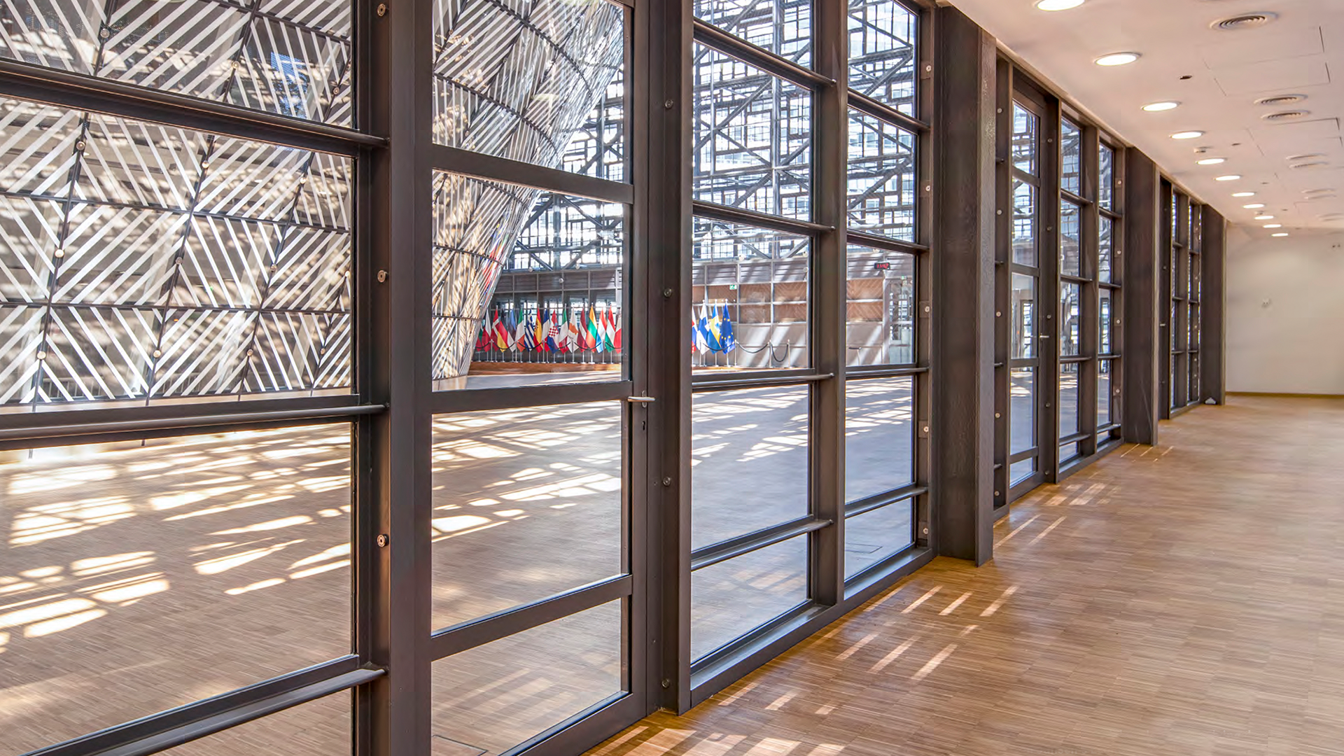 View through glass walls into a modern building with a display of international flags inside, sunlight casting shadows on the wood floor, and a curved architectural structure visible through the windows.