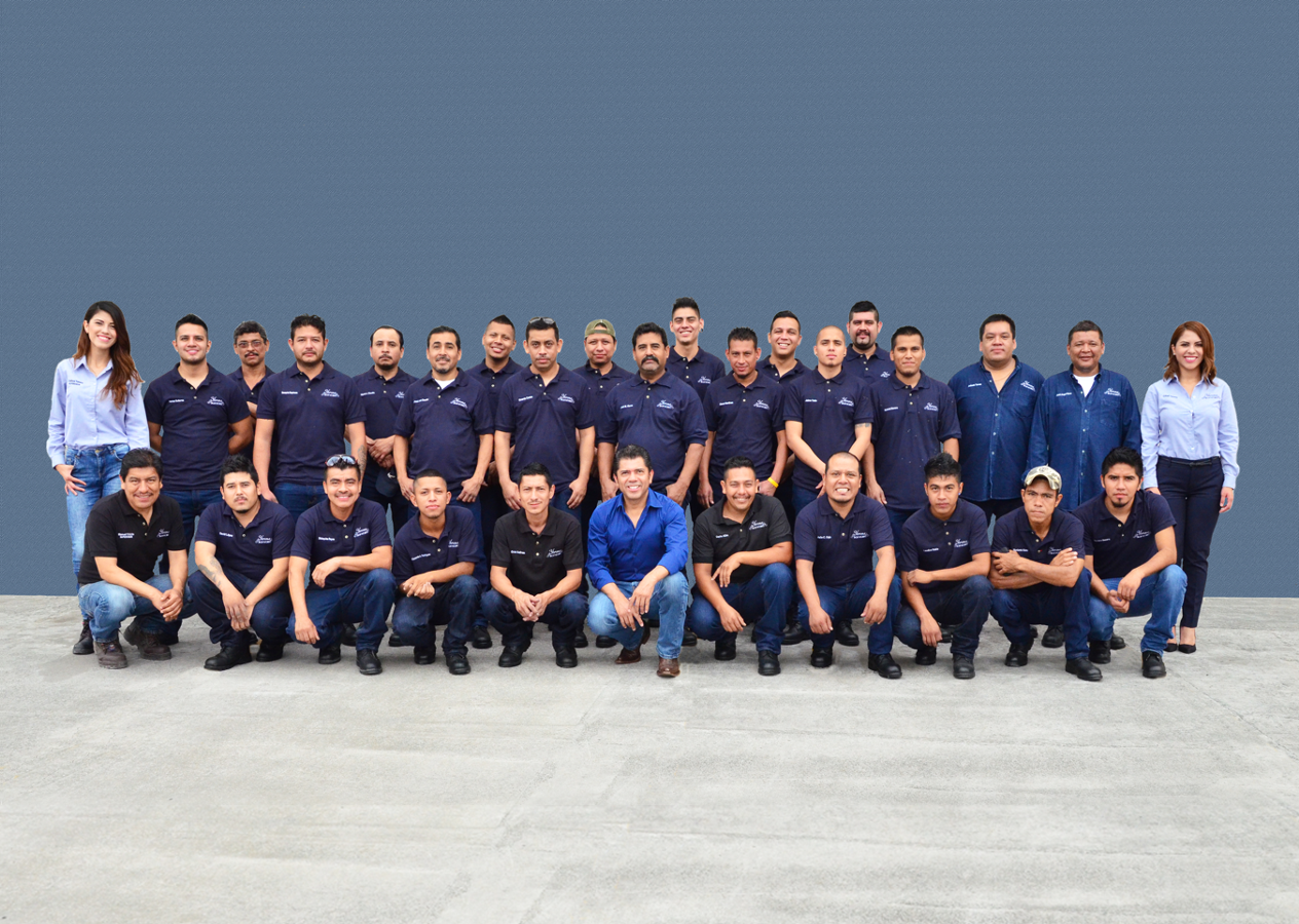 Group photo of 22 people, mostly men, standing and kneeling in front of a plain blue wall, wearing matching dark blue shirts with logos, two women at the ends wearing light blue shirts, on a concrete ground.