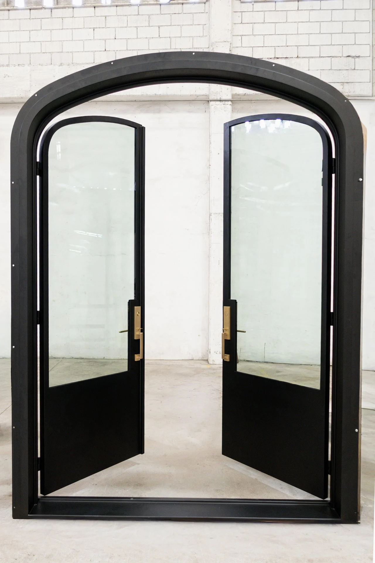 Double glass doors with black frames and brass handles, set in a black metal frame with an arched top, in an industrial space with white brick walls.