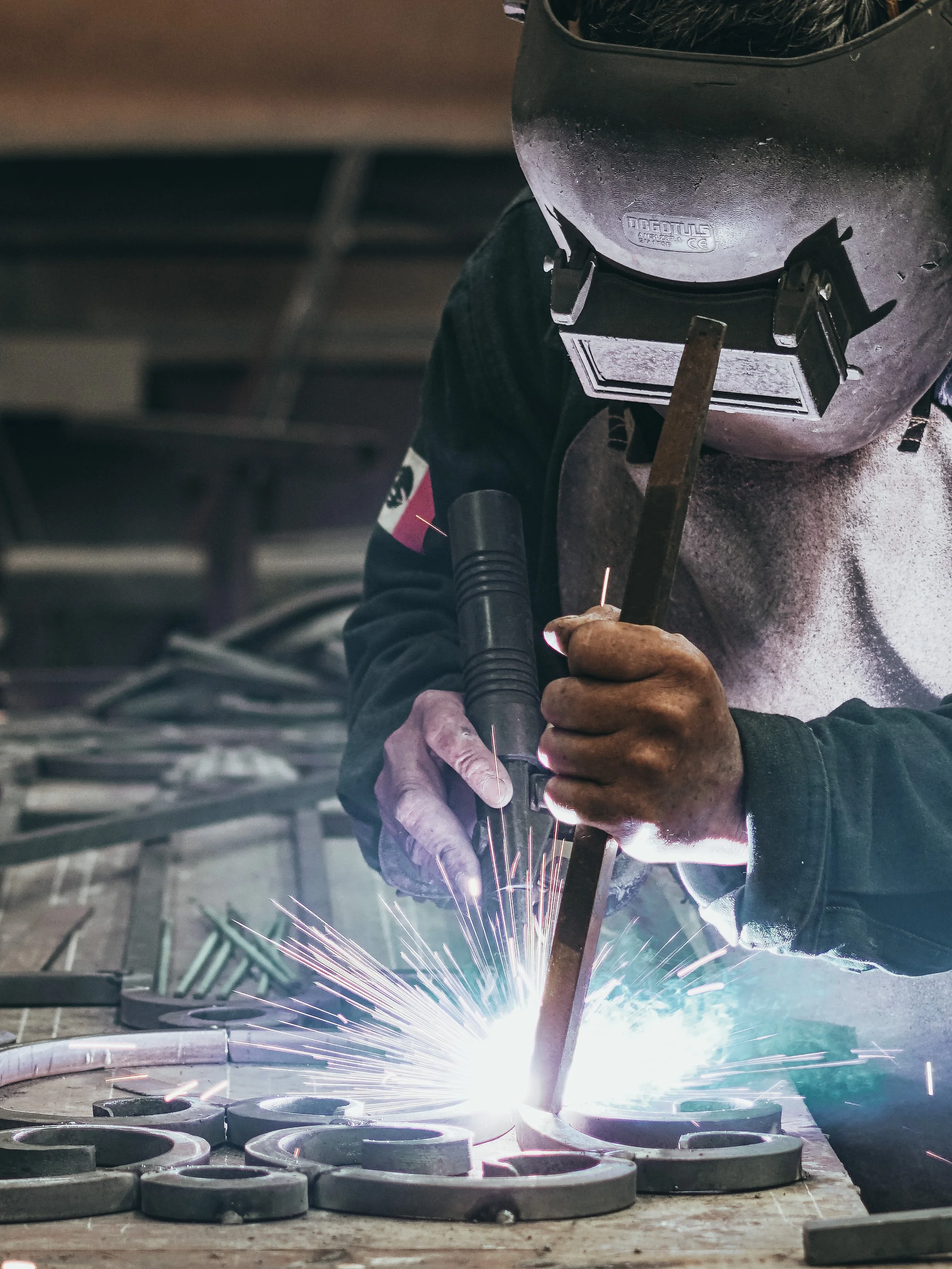 A welder wearing protective gear, including a welding helmet and gloves, working on a metal project, with sparks flying from the welding process in an industrial workshop.