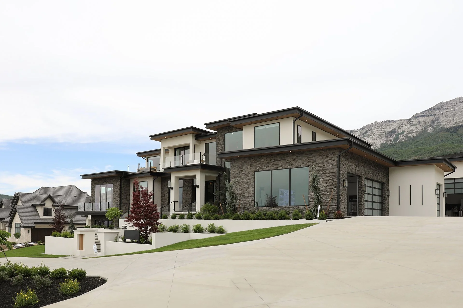 Modern multi-story house with stone and white exterior walls, large windows, balconies, and a sloped driveway leading to a garage, with landscaped yard and mountainous background.
