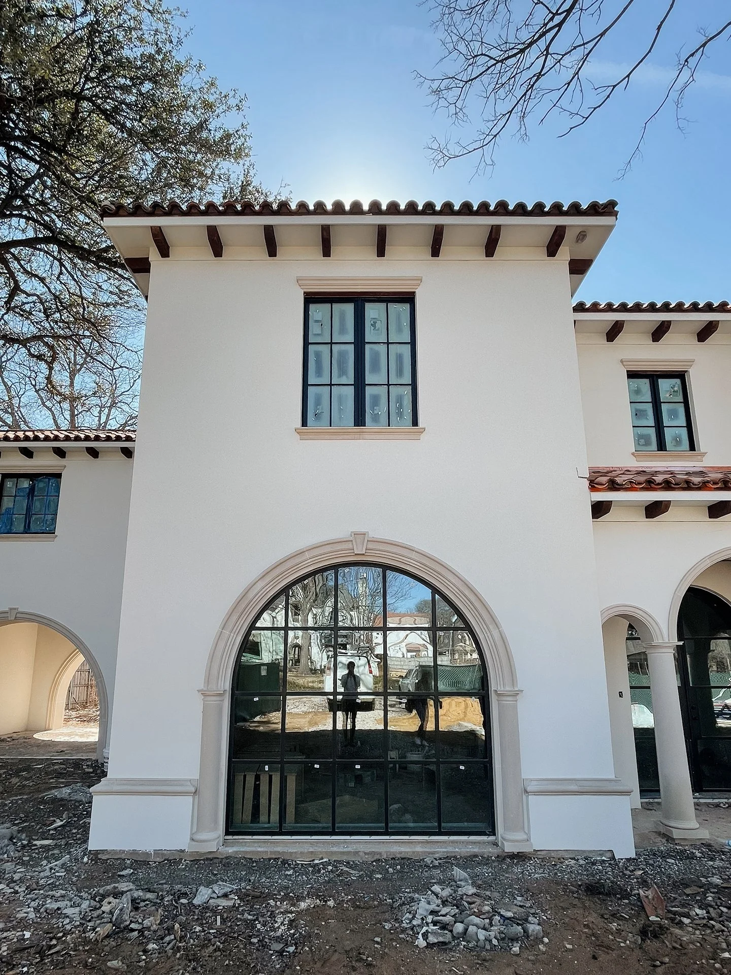 The front view of a building under construction or renovation with a large arched window reflecting trees and the sky, surrounded by a white facade and decorative columns, with a construction yard below and trees visible in the background.